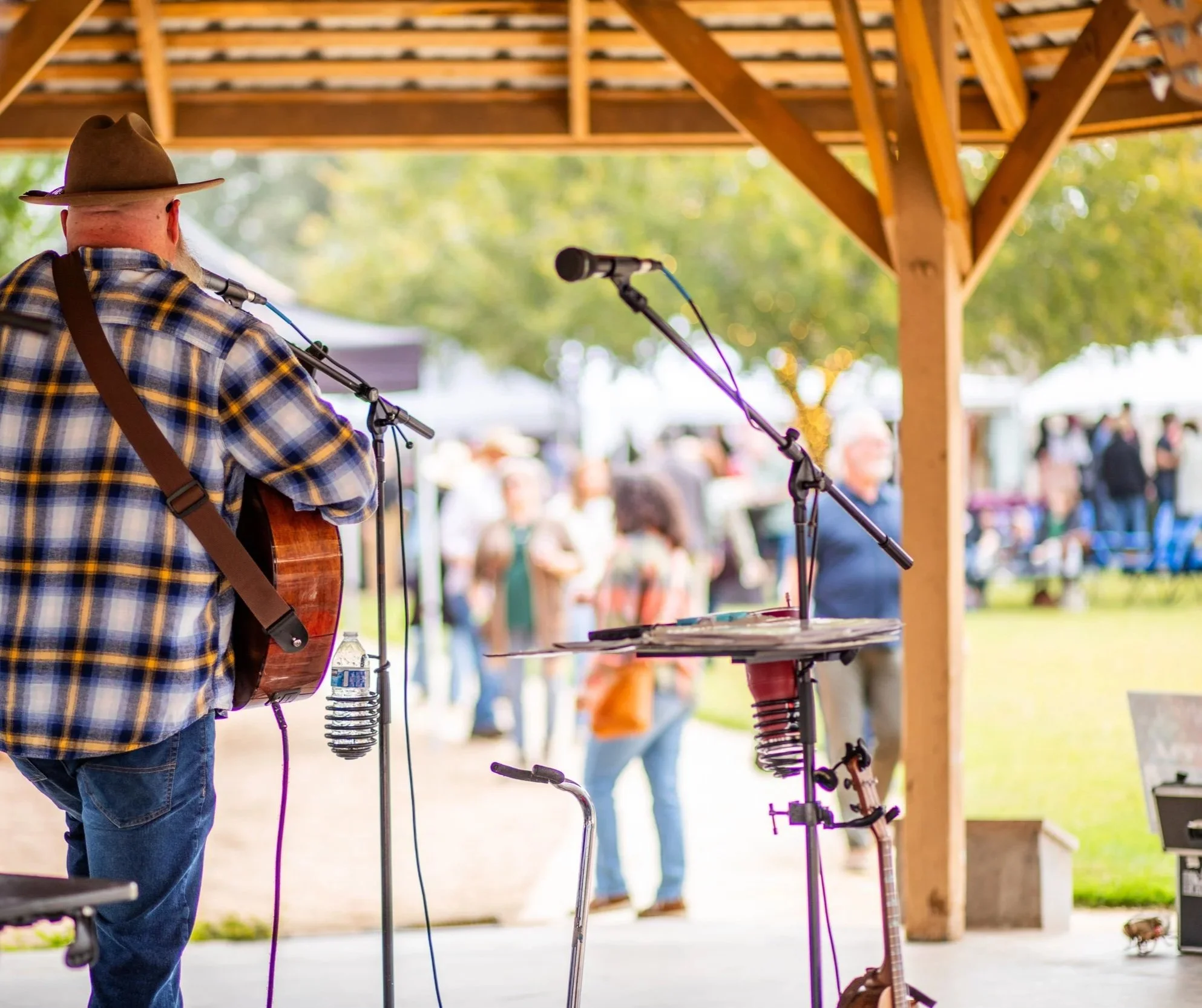 A musician with a beard and a wide-brimmed hat playing guitar on an outdoor stage under a wooden pavilion during a daytime event, with people and trees in the background.