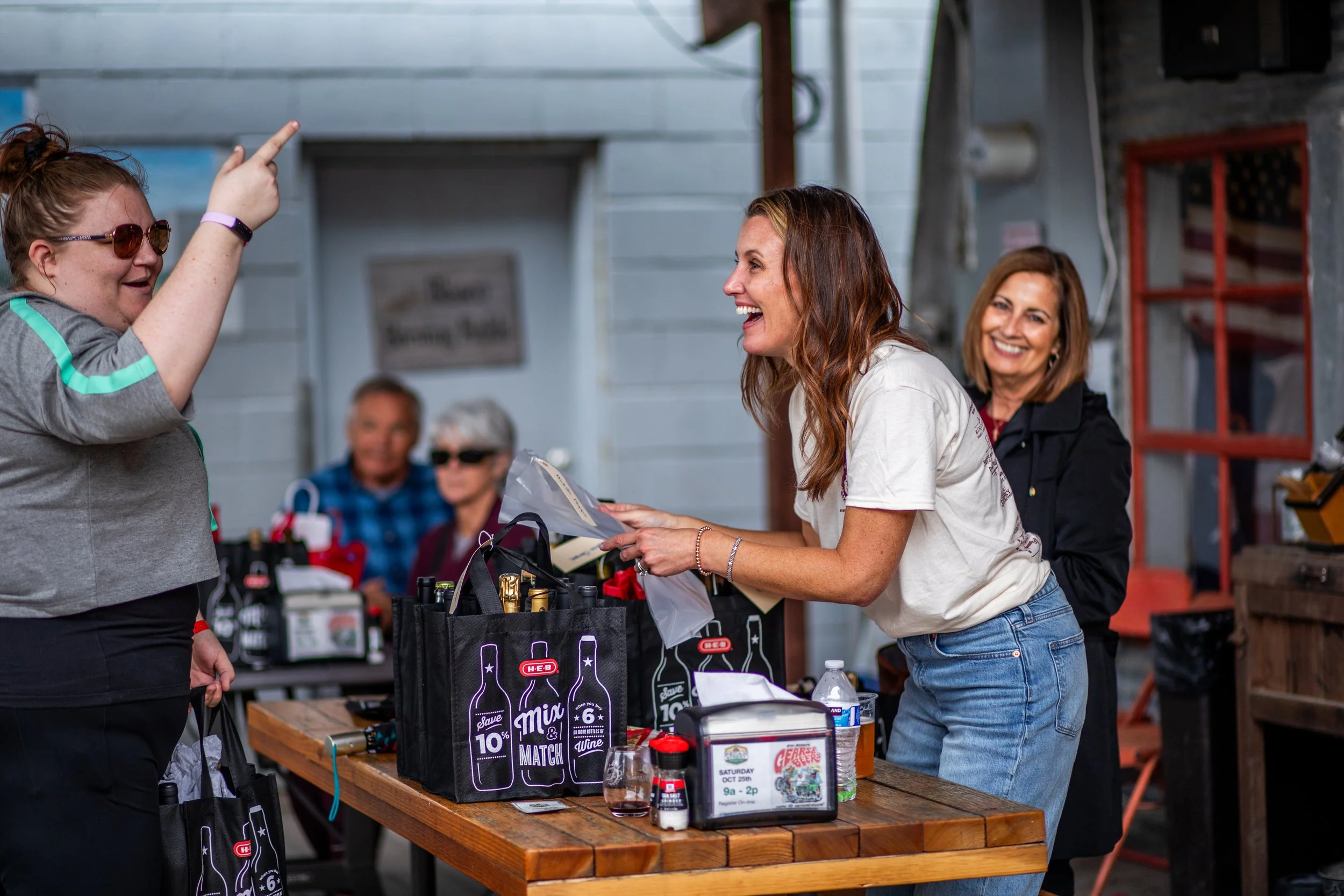 A woman with long red hair, wearing a white t-shirt and jeans, is smiling and interacting with another woman who is pointing and wearing sunglasses and a gray shirt. They are at a table in a rustic indoor setting, likely at a market or fair, with various bottles, signs, and bags on the table, and other people in the background.