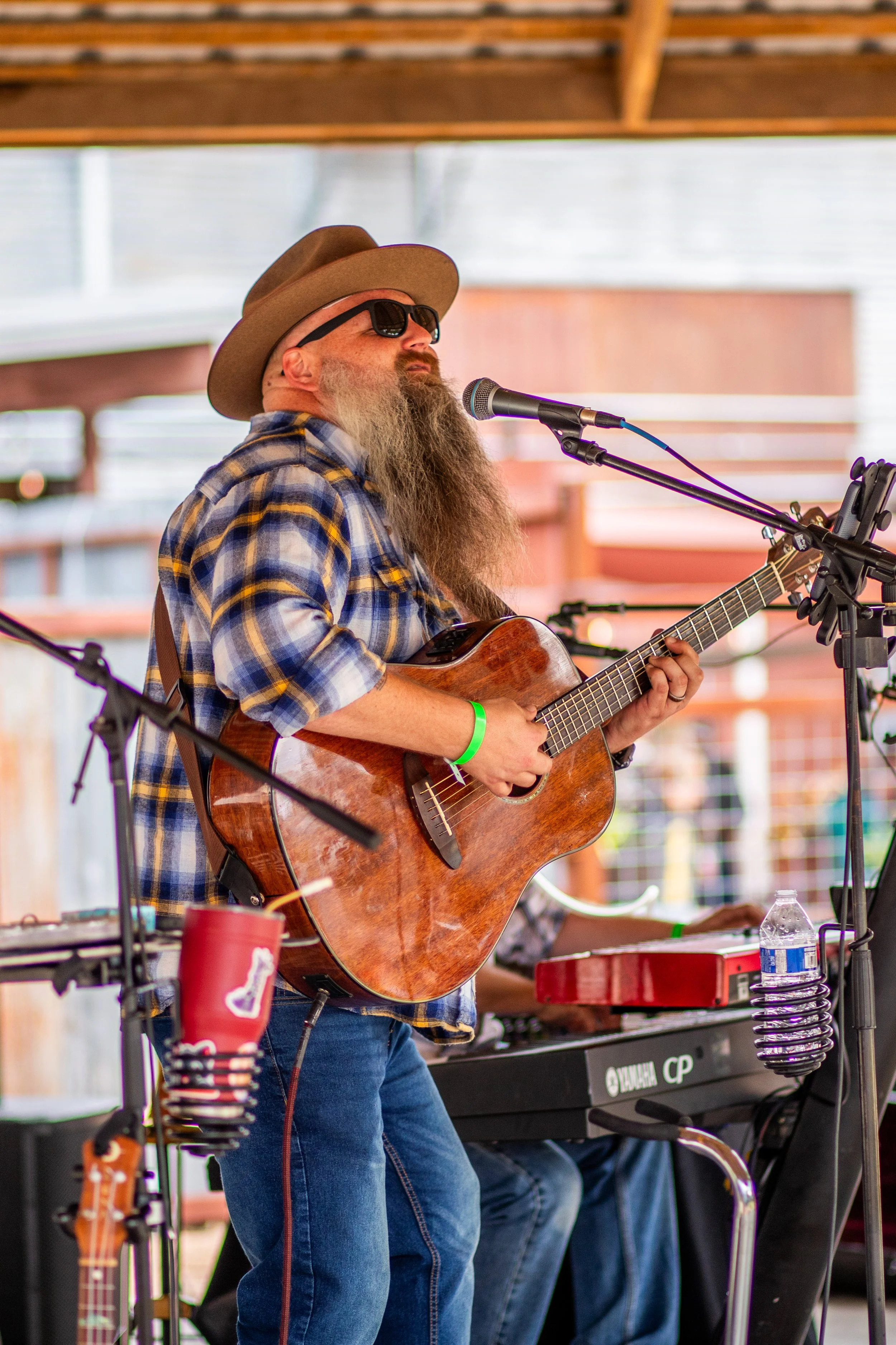 Man with a long beard, sunglasses, and a brown hat plays an acoustic guitar and sings into a microphone at an outdoor event.