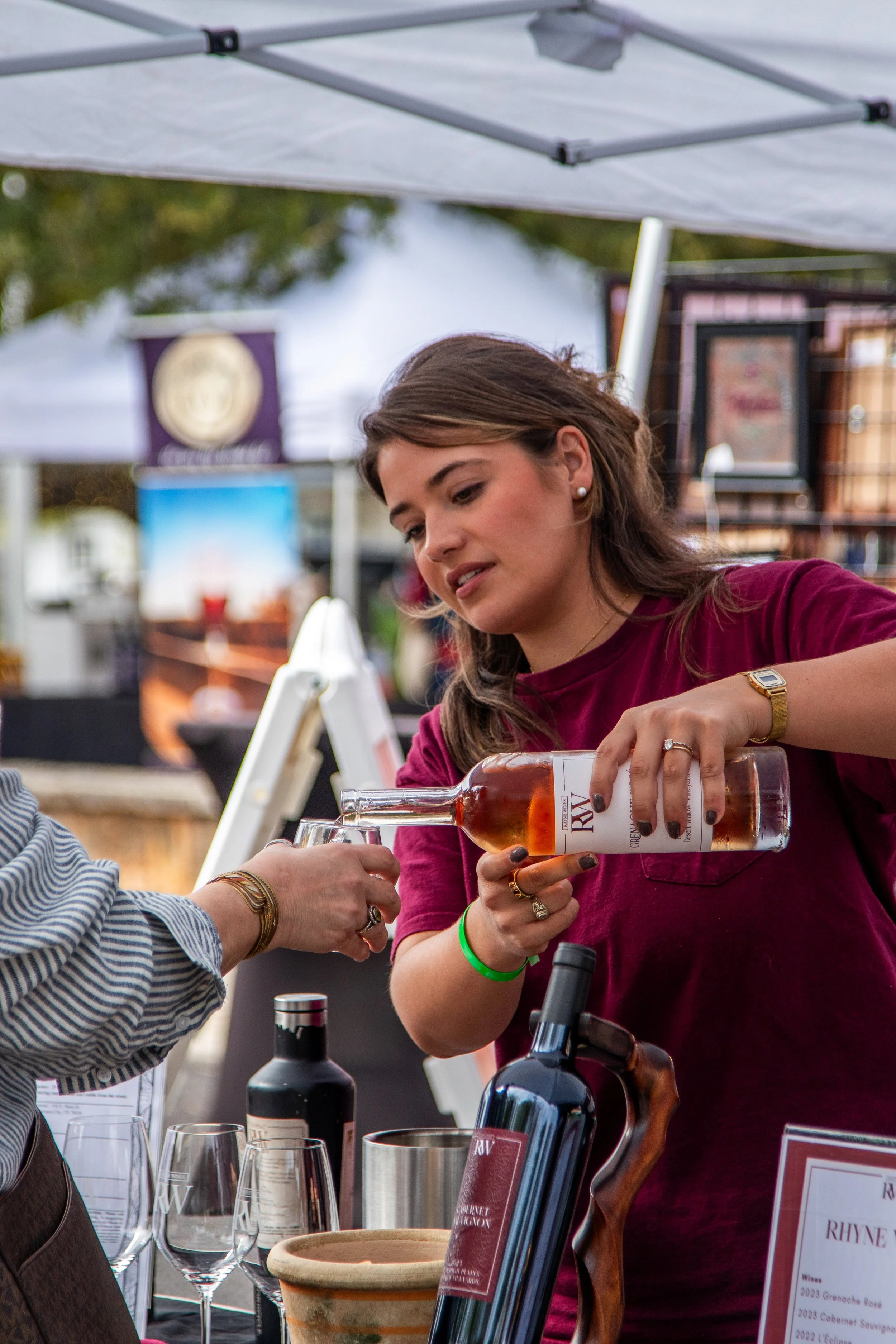 Woman in maroon shirt pouring wine at outdoor wine tasting event, with tables, wine bottles, glasses, and tasting notes in the background.