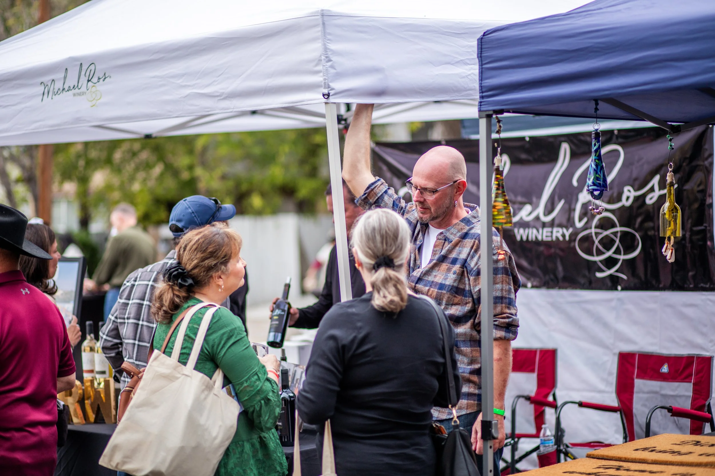 People gathered at an outdoor event around a vendor booth with a man in glasses and a plaid shirt talking to others under a white canopy with a winery logo.