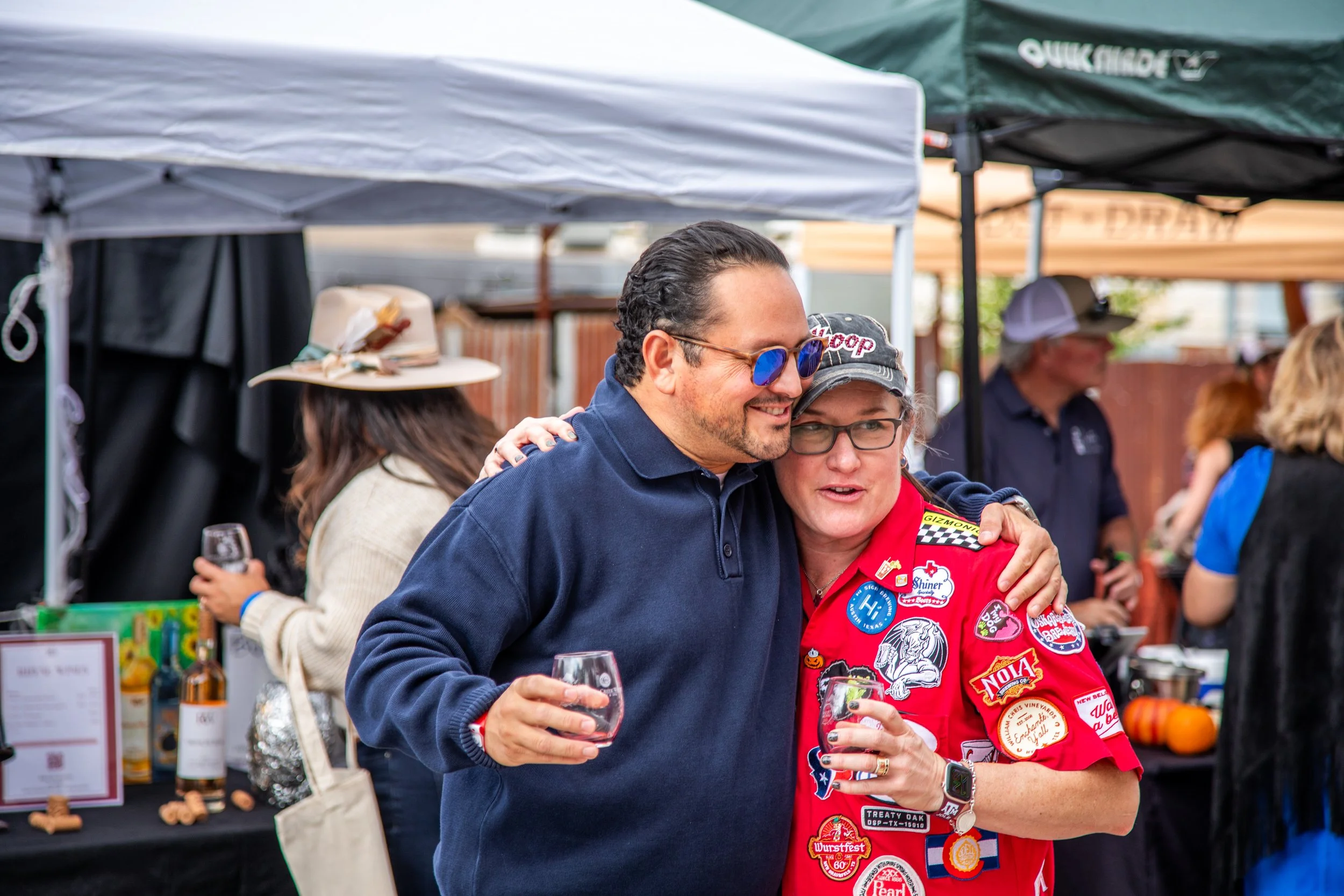 Two people hugging at an outdoor event with tents and booths in the background. One person is wearing a navy blue shirt and sunglasses, and the other is wearing a red racing jacket with various patches. They are holding glasses of wine.