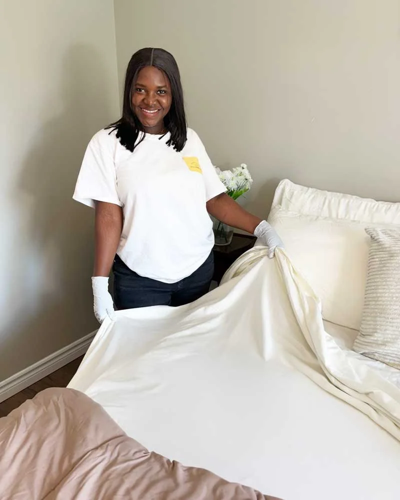 A woman with dark hair, wearing a white t-shirt and gloves, making  a bed with white sheets in a bedroom.