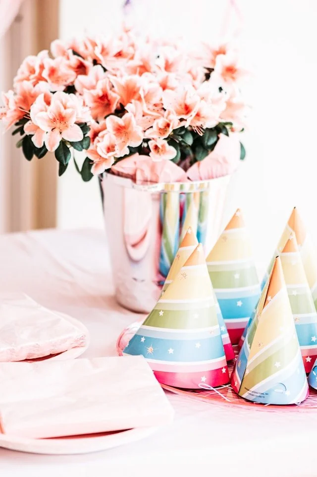 A birthday table setting with a bouquet of pink flowers in a shiny silver vase, pink napkins, and colorful party hats with pastel rainbow stripes.