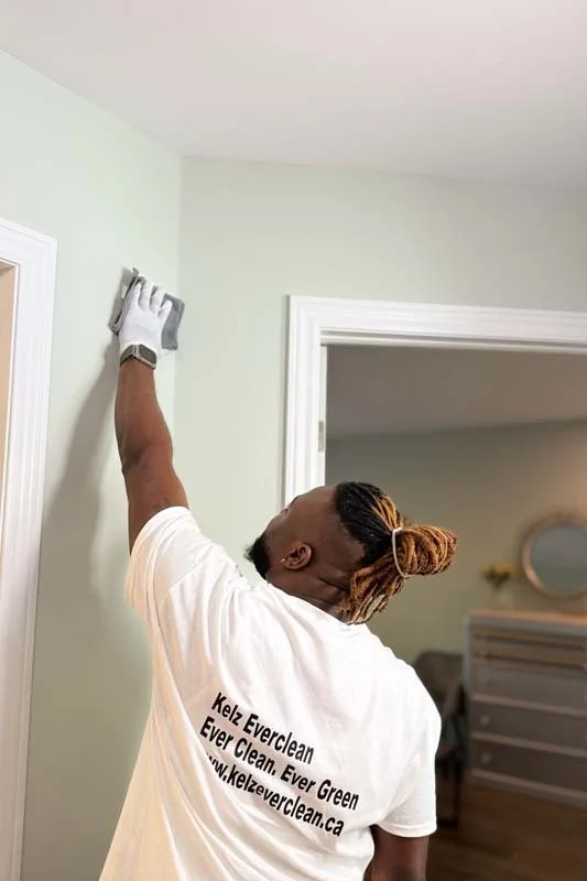 A man cleaning a light green wall in a room, wearing a white t-shirt with black text and a white cleaning glove on his right hand, reaching up to wipe the wall.
