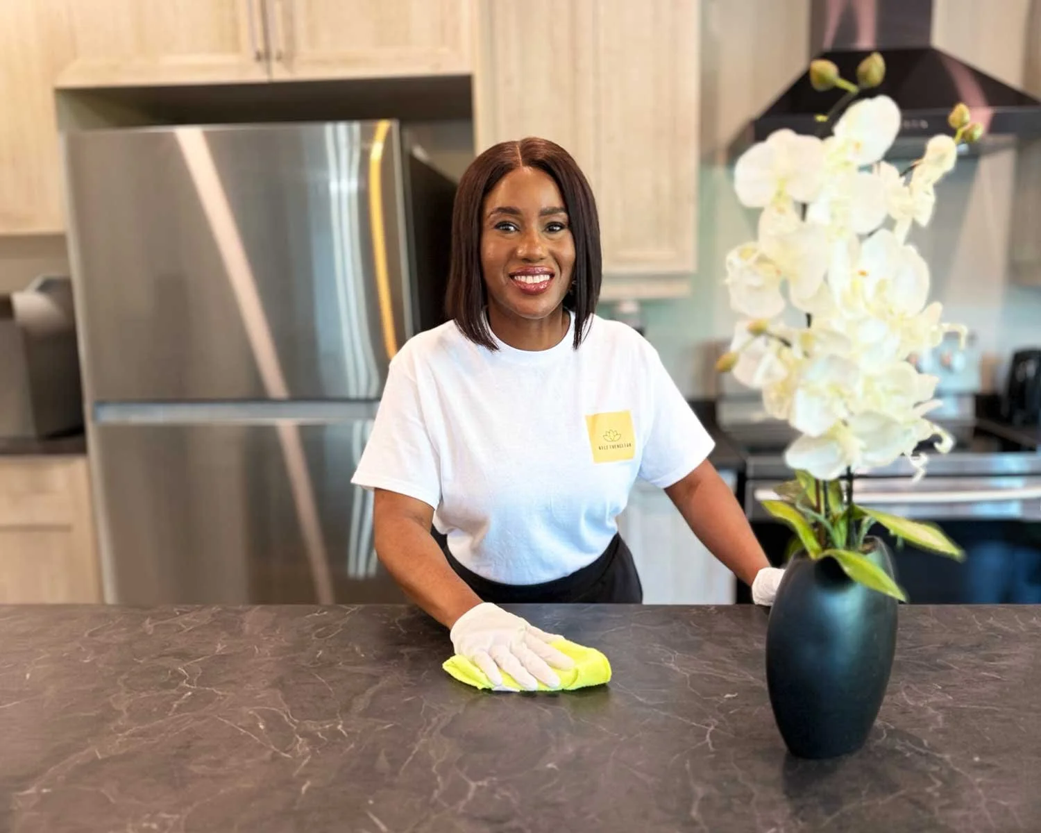 A woman with shoulder-length dark hair smiling while cleaning a kitchen counter with a yellow cloth. She is wearing a white T-shirt with a yellow logo and gloves, standing next to a black vase with white orchids.