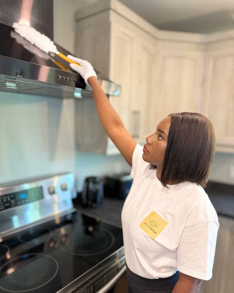 Woman cleaning a kitchen range hood with a sponge, wearing gloves, in a home kitchen.