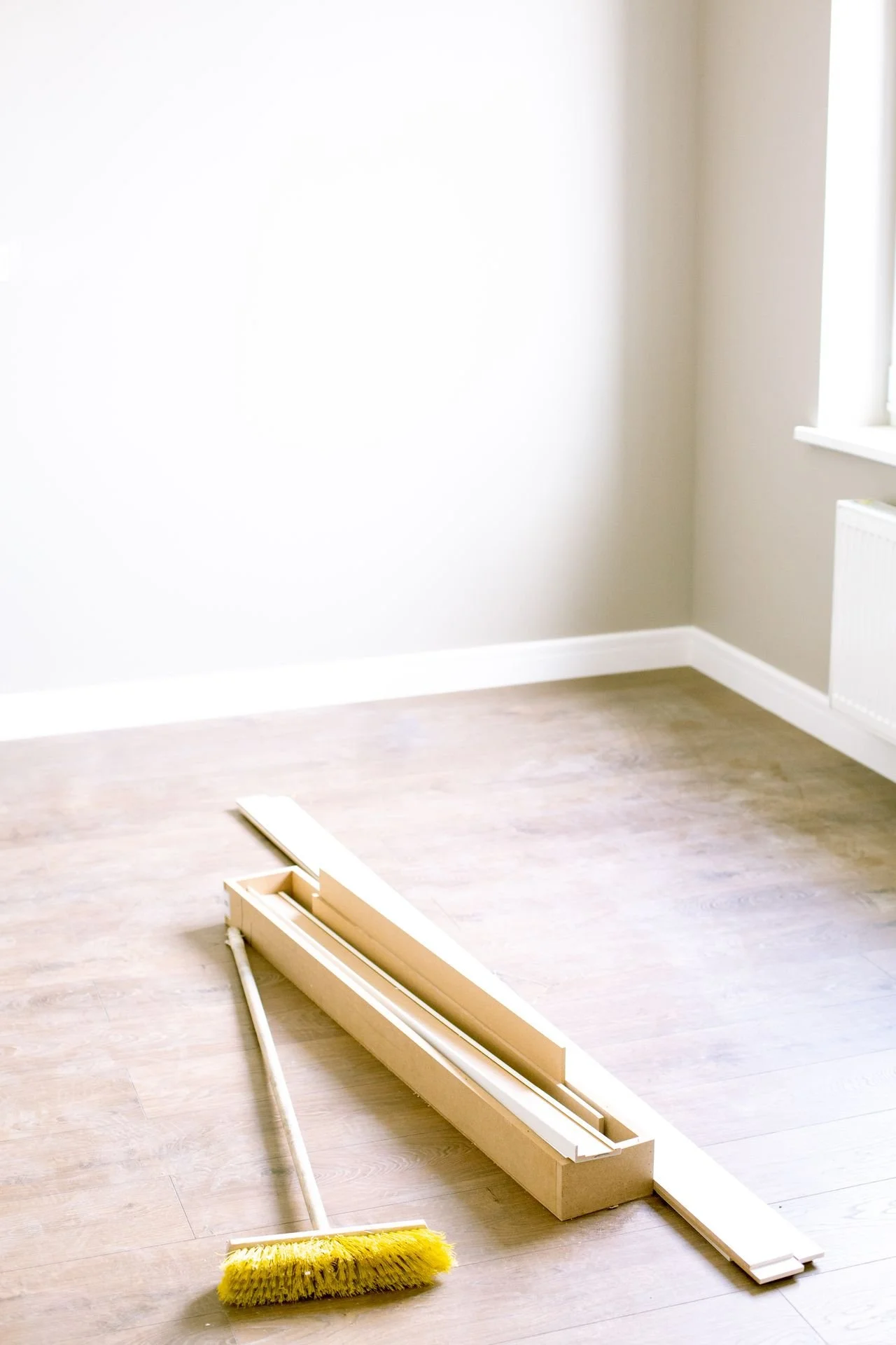 Empty room with light-colored wooden floor, a broom, and a partially assembled piece of furniture on the floor, next to a window and a radiator.