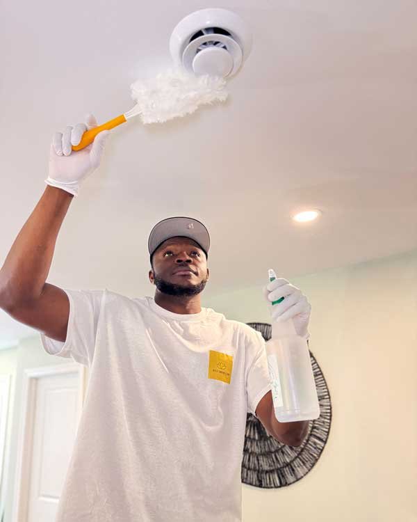 A man wearing a white t-shirt, gloves, and a baseball cap is cleaning a ceiling fan with a duster while holding a spray bottle.