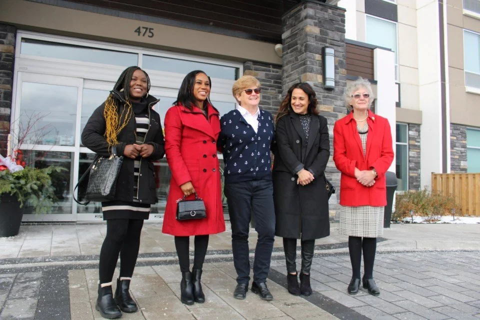 Five women standing side by side outside a modern building with glass doors and stone accents.