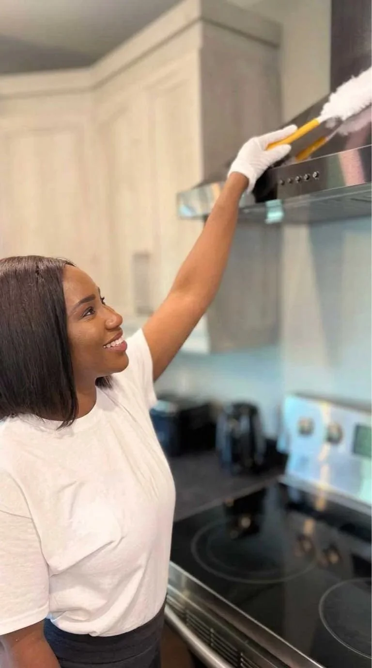 A woman with a white T-shirt and gloves cleaning a kitchen range hood with a duster.