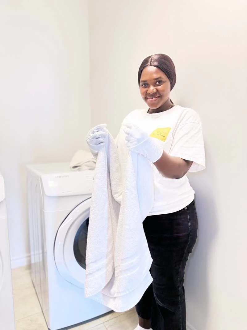 A woman with short dark hair wearing a white t-shirt with a yellow logo, white gloves, and black pants, standing next to a washing machine, holding a white towel.