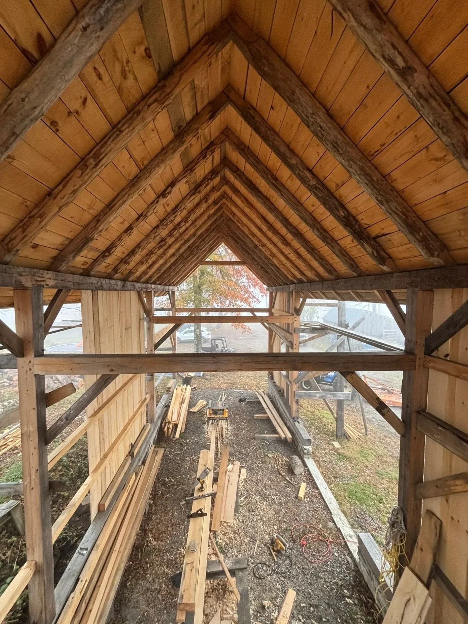 Construction site with wooden framework, beams, and planks forming an arched roof, with tools and materials scattered on the ground. Construction of a timber frame barn with Hewn beams and tradtional construction techniques.