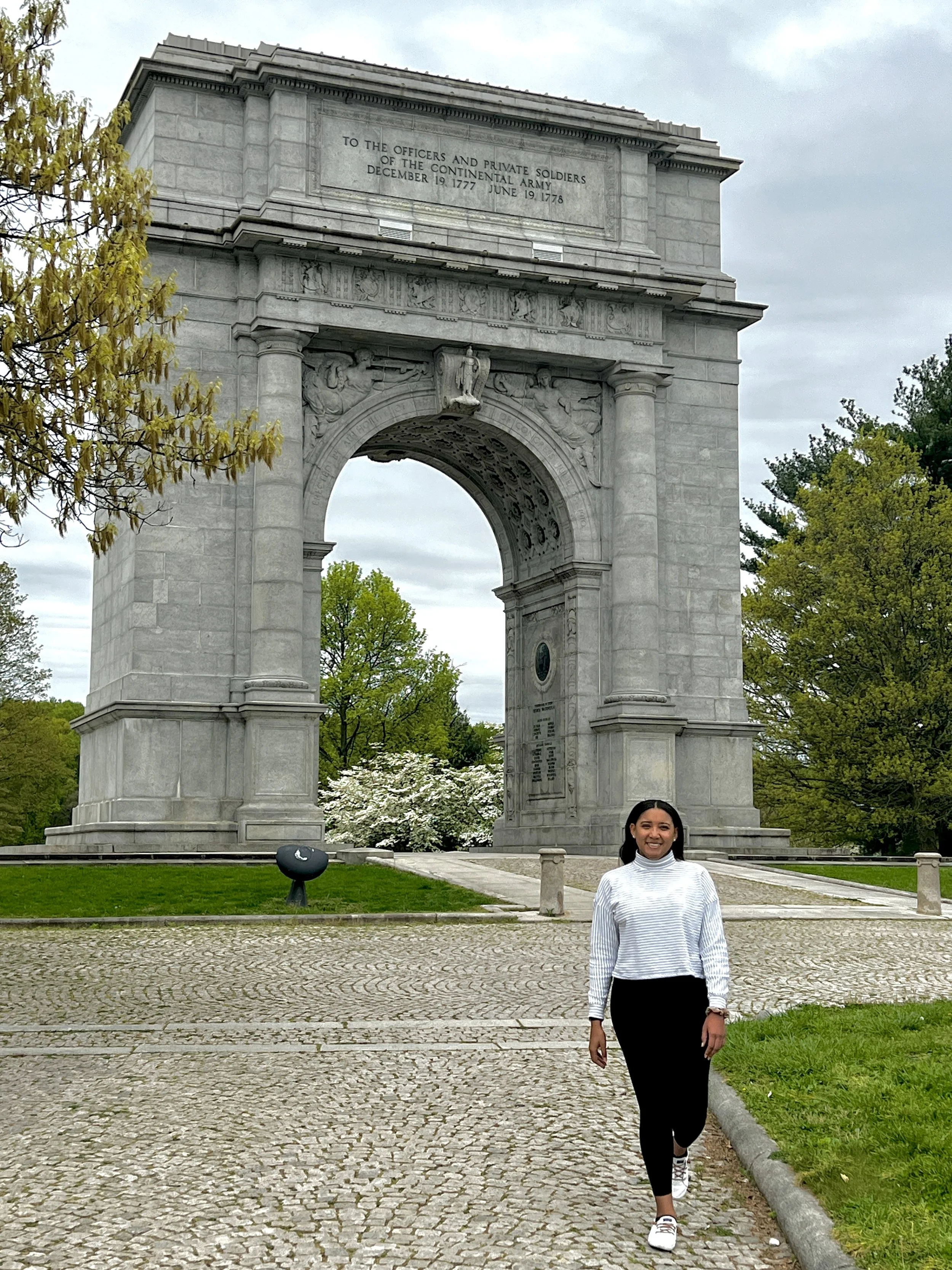 Woman walking in front of a large stone arch monument, smiling, wearing a white and gray striped sweater, black pants, and sneakers, surrounded by trees with green leaves.