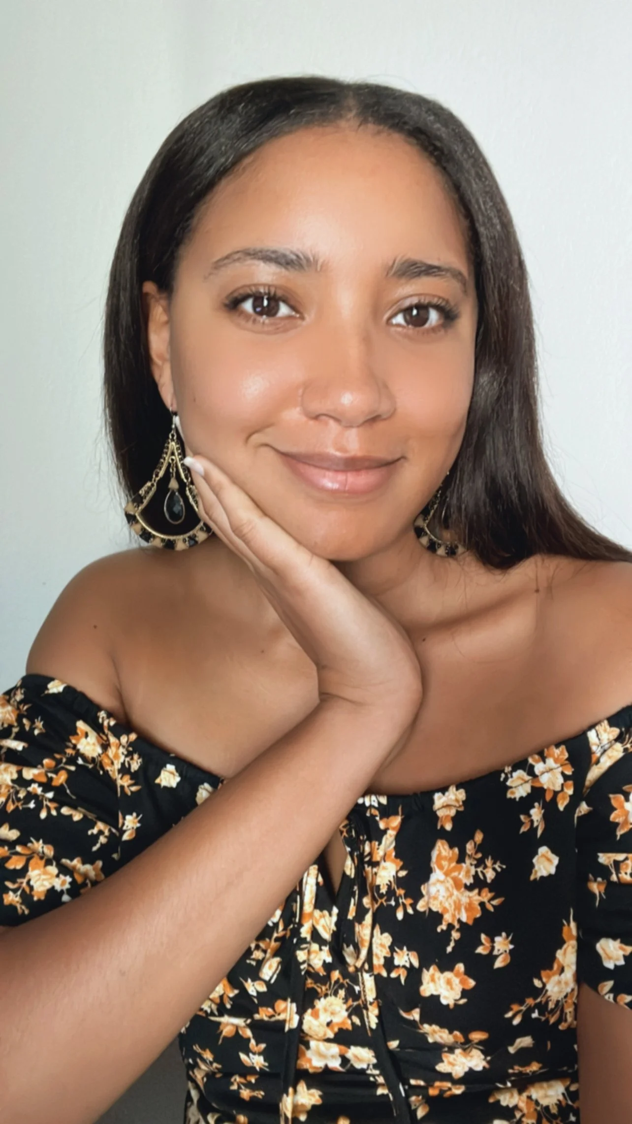 A young woman with long dark hair, wearing floral off-the-shoulder top and large earrings, smiling with her hand resting on her chin, against a plain light-colored background.