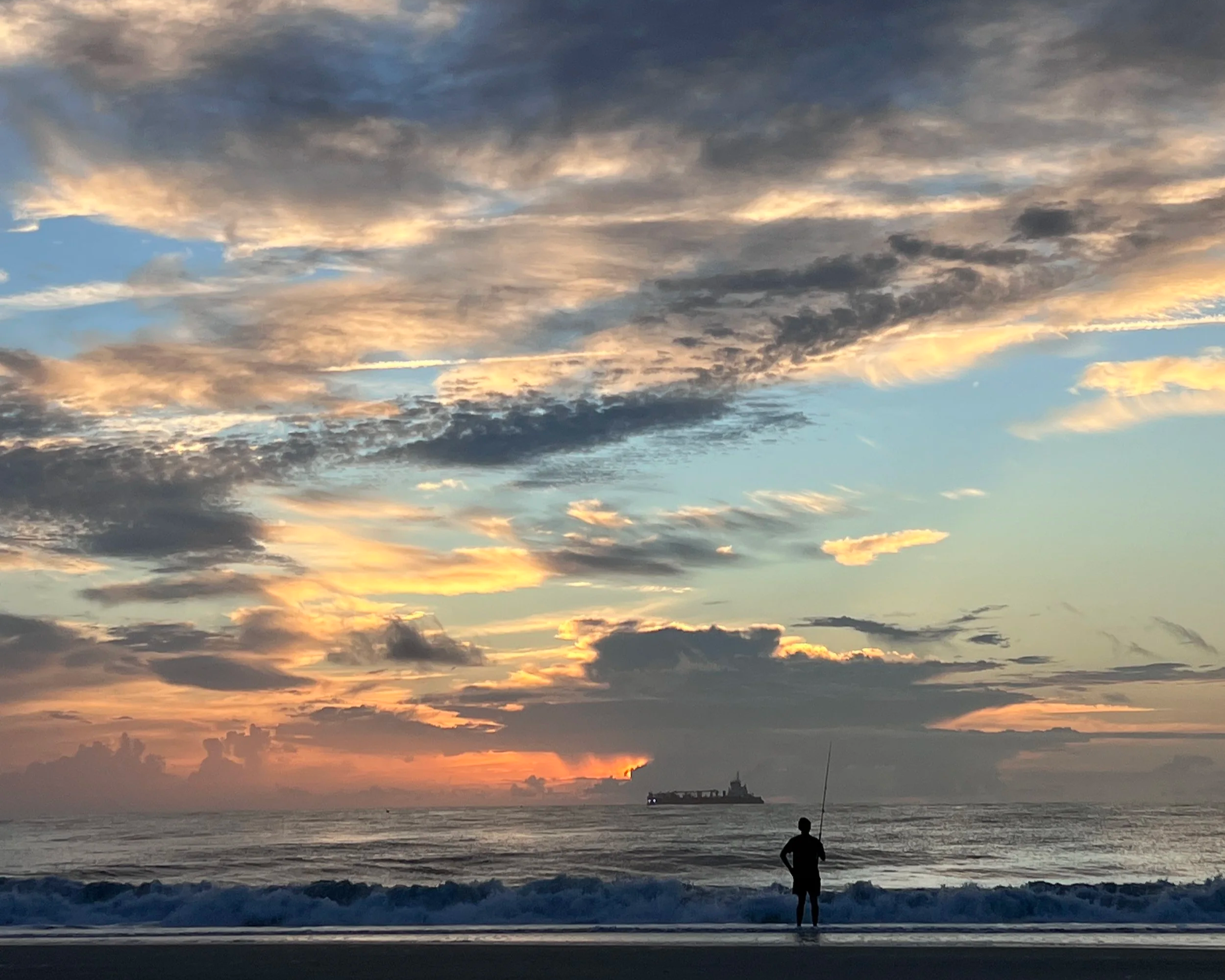 A sunset over the ocean with partially cloudy sky, a boat on the water, and a person standing on the beach fishing.