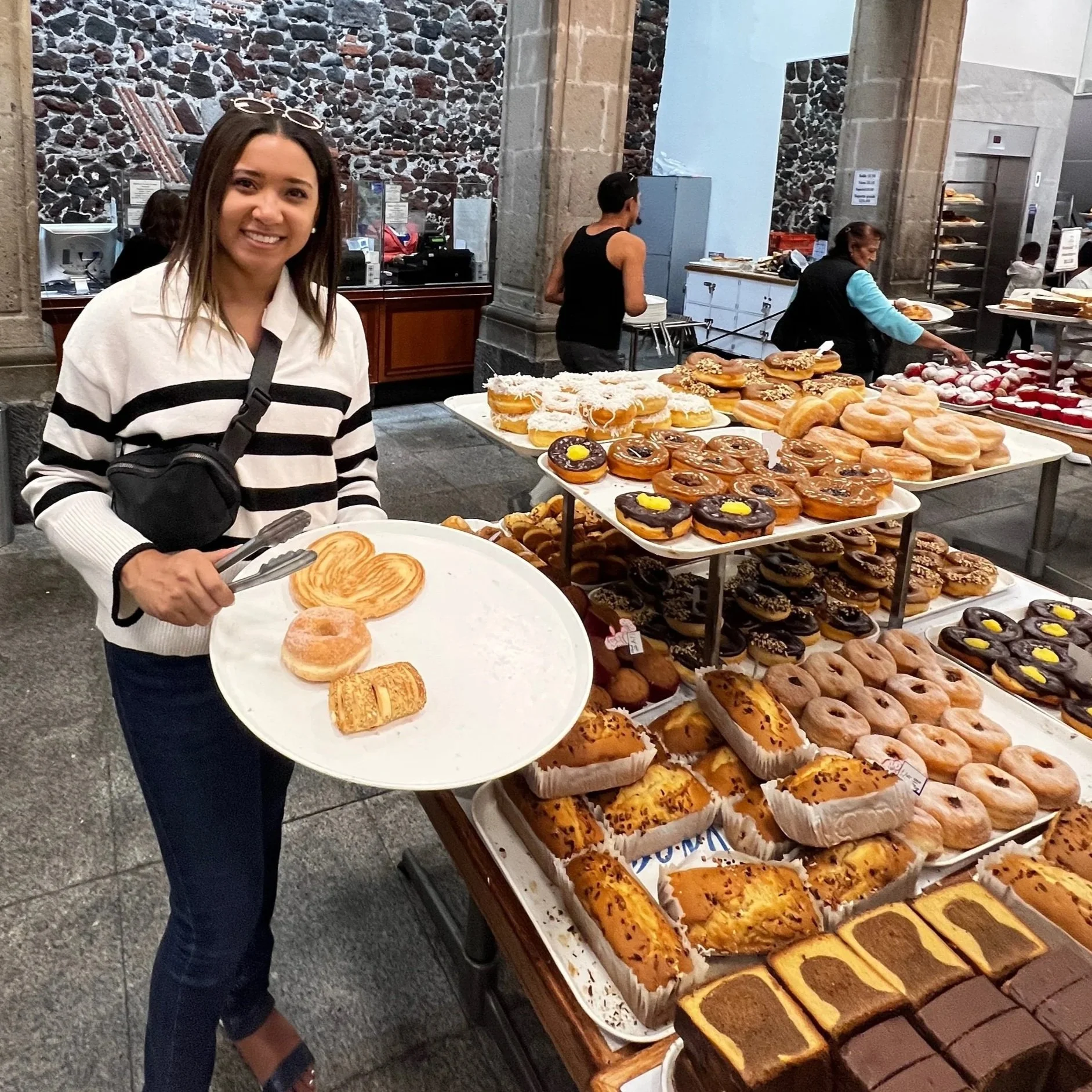 A woman smiling and holding tongs stands next to a table with a large variety of baked goods, including donuts, pastries, and cakes, in a bakery or cafe setting.