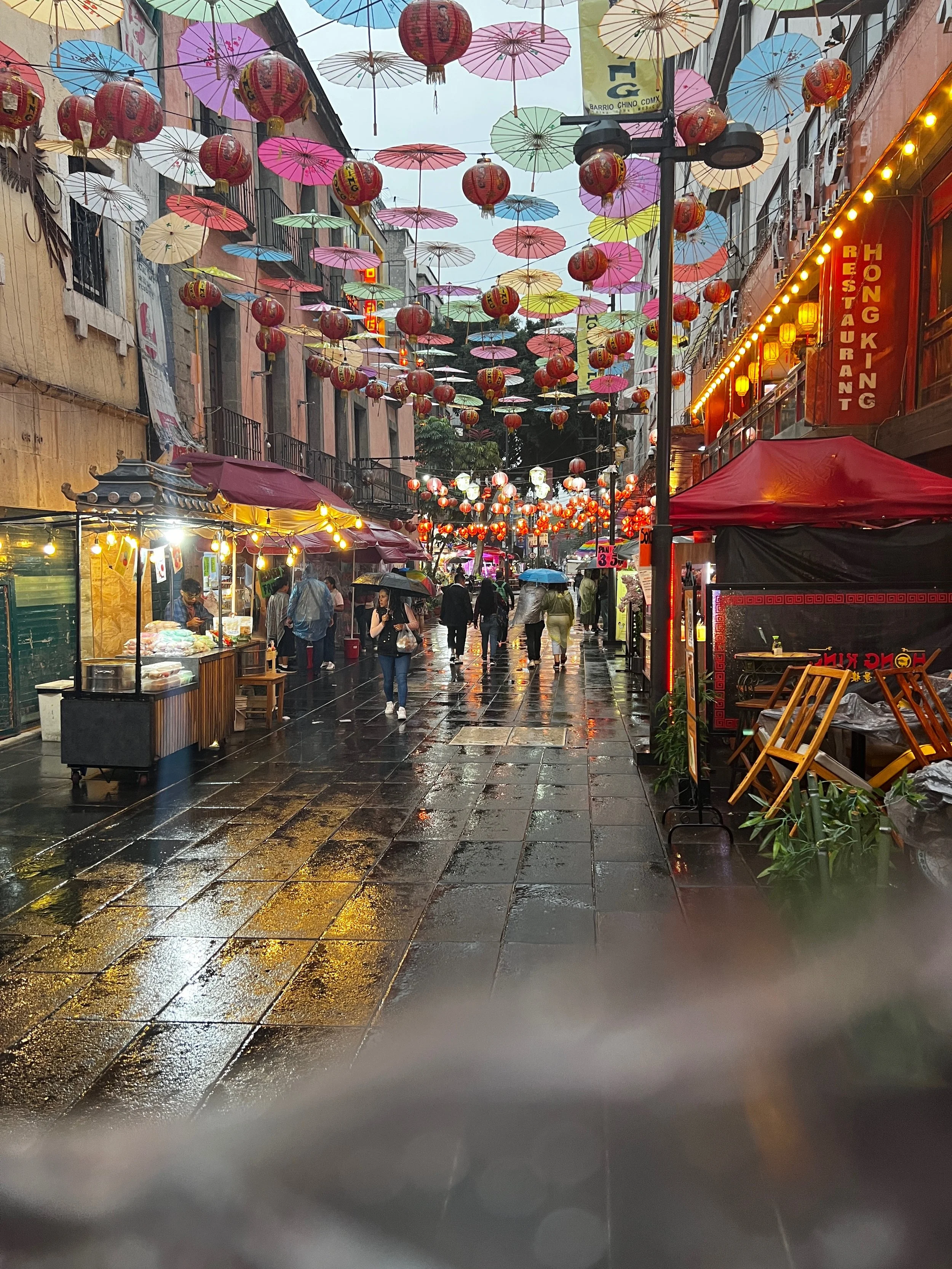 A vibrant street scene after rain, decorated with colorful umbrellas and red lanterns hanging above. People with umbrellas walk along a wet sidewalk, shops with illuminated signs line both sides of the street.