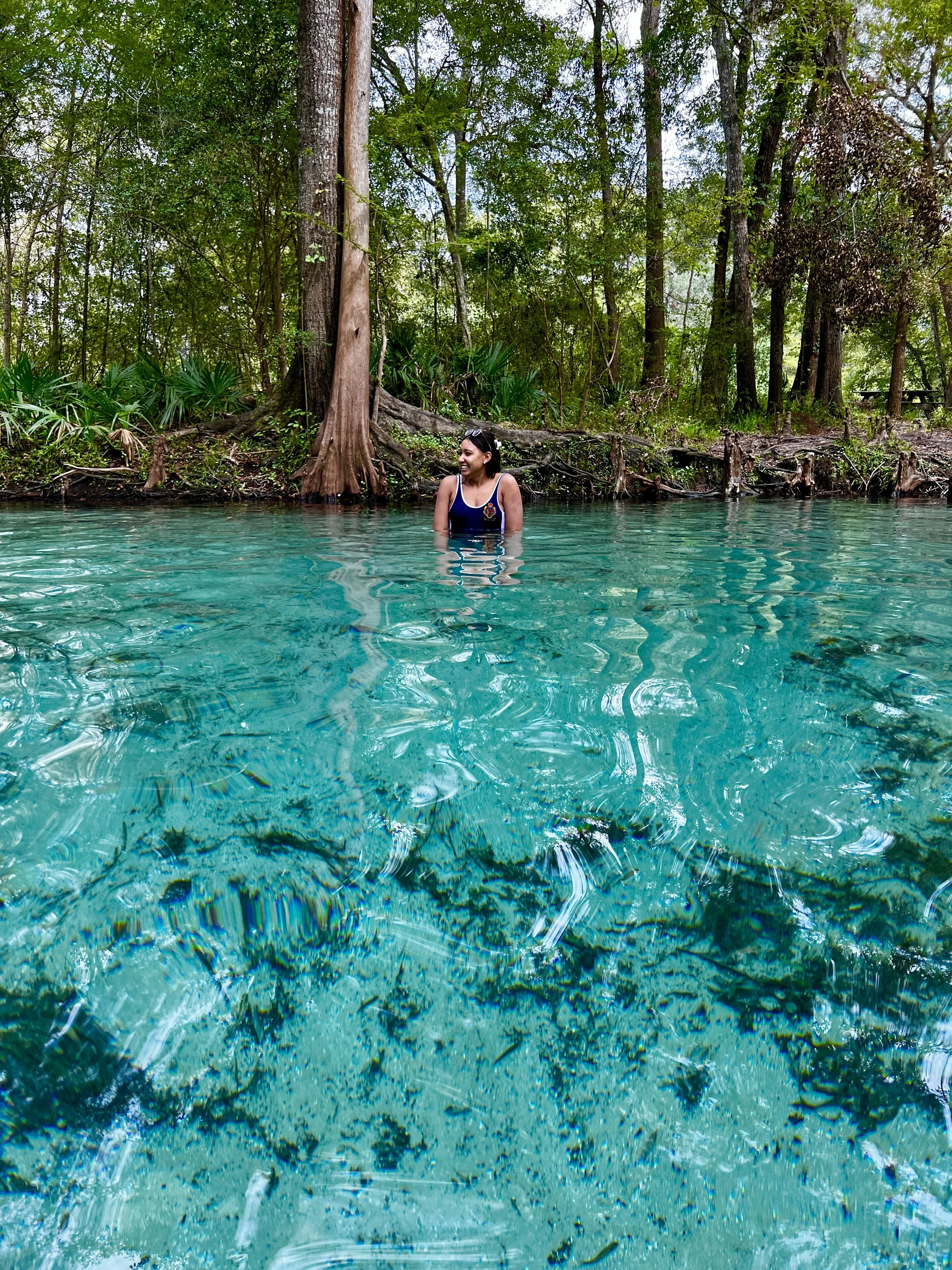 A woman wearing a swimsuit and sunglasses enjoying herself in a natural freshwater pool surrounded by trees and lush greenery.