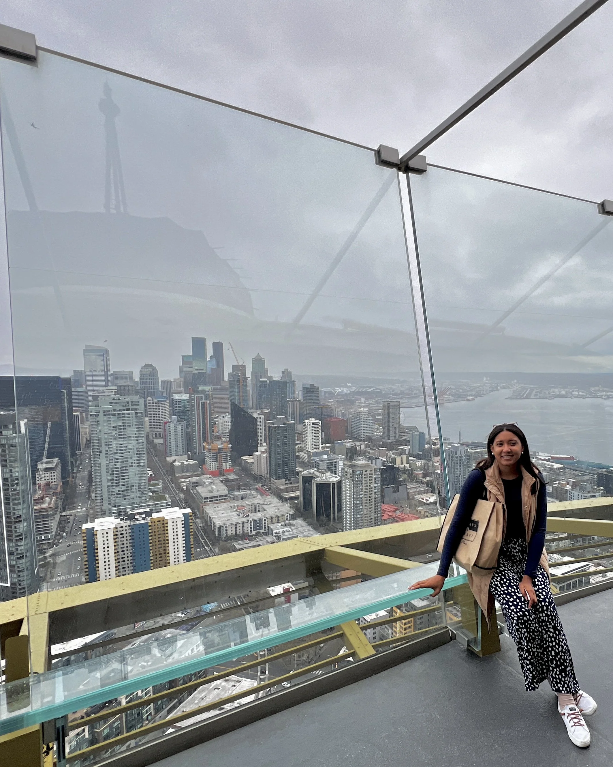A woman standing on an observation deck of a tall building with a city skyline and a river in the background. The deck has glass panels and metal railings.