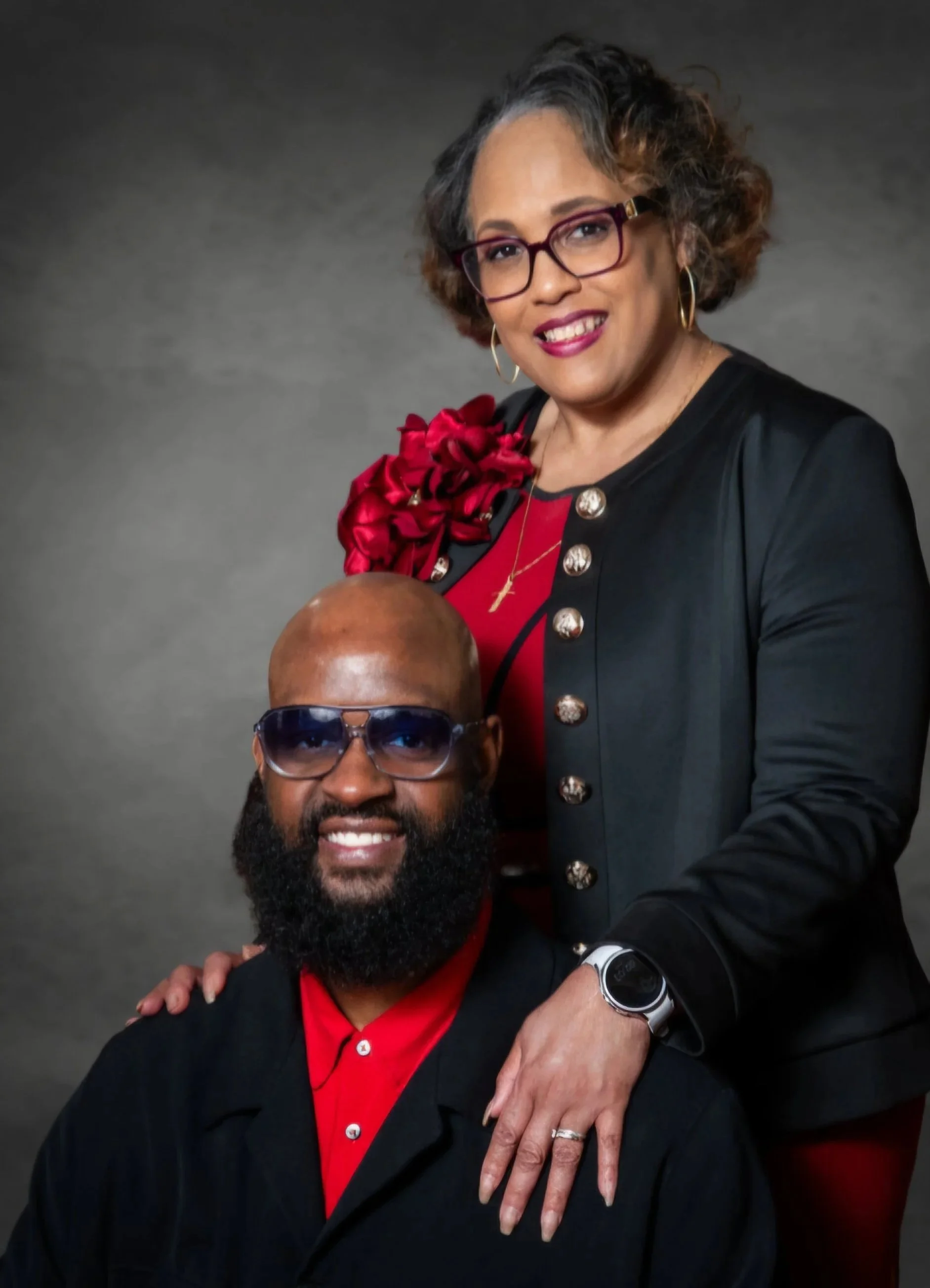 A woman with glasses, short curly hair, and hoop earrings, standing behind a man with glasses, a beard, and a bald head. She is smiling and wearing a black jacket with gold buttons and a red top with a decorative red flower on her left shoulder. The man is wearing a black jacket over a red shirt, has a wristwatch, and is smiling with his face partially visible. The background is a plain, dark gray backdrop.