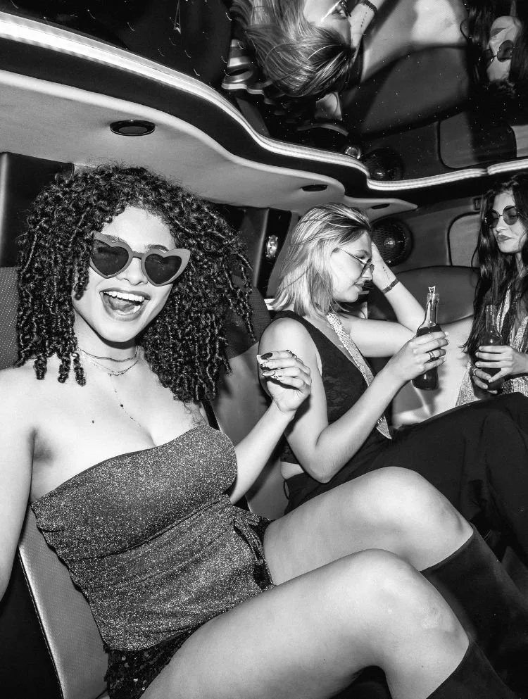 Drei Frauen sitzen in einer Limousine, trinken und feiern, eine trägt Herz-Aussicht sunglasses, alle sehen entspannt und fröhlich aus.