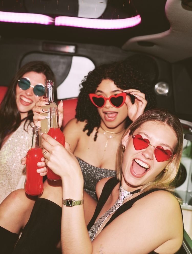 Drei Frauen feiern in einem Auto, tragen Herzbrillen, halten Getränke und lächeln fröhlich.