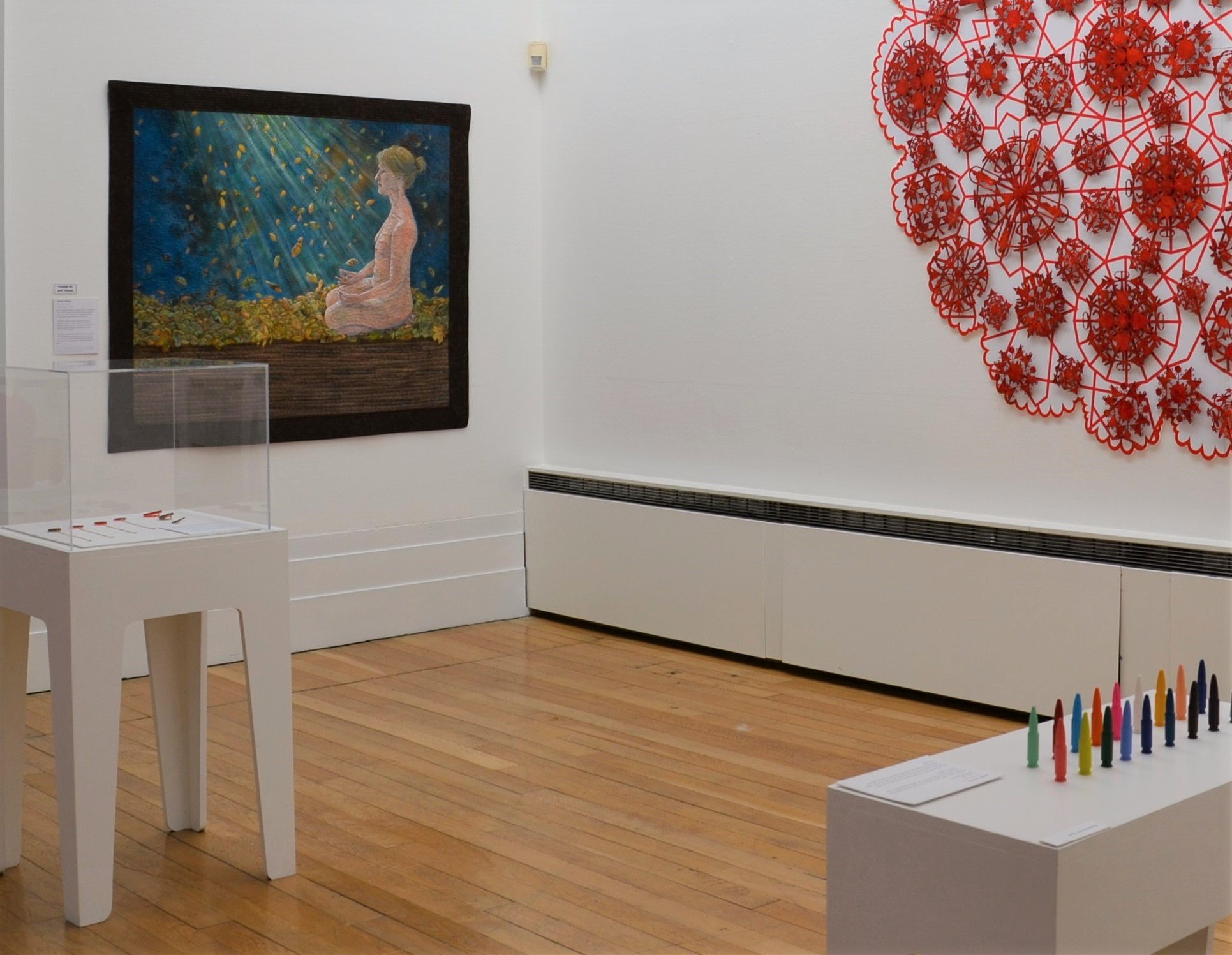 Art gallery interior with a painting of a seated figure surrounded by light, a red lace-like wall sculpture, a display case on a white stand, and colorful cone-shaped objects on a bench.