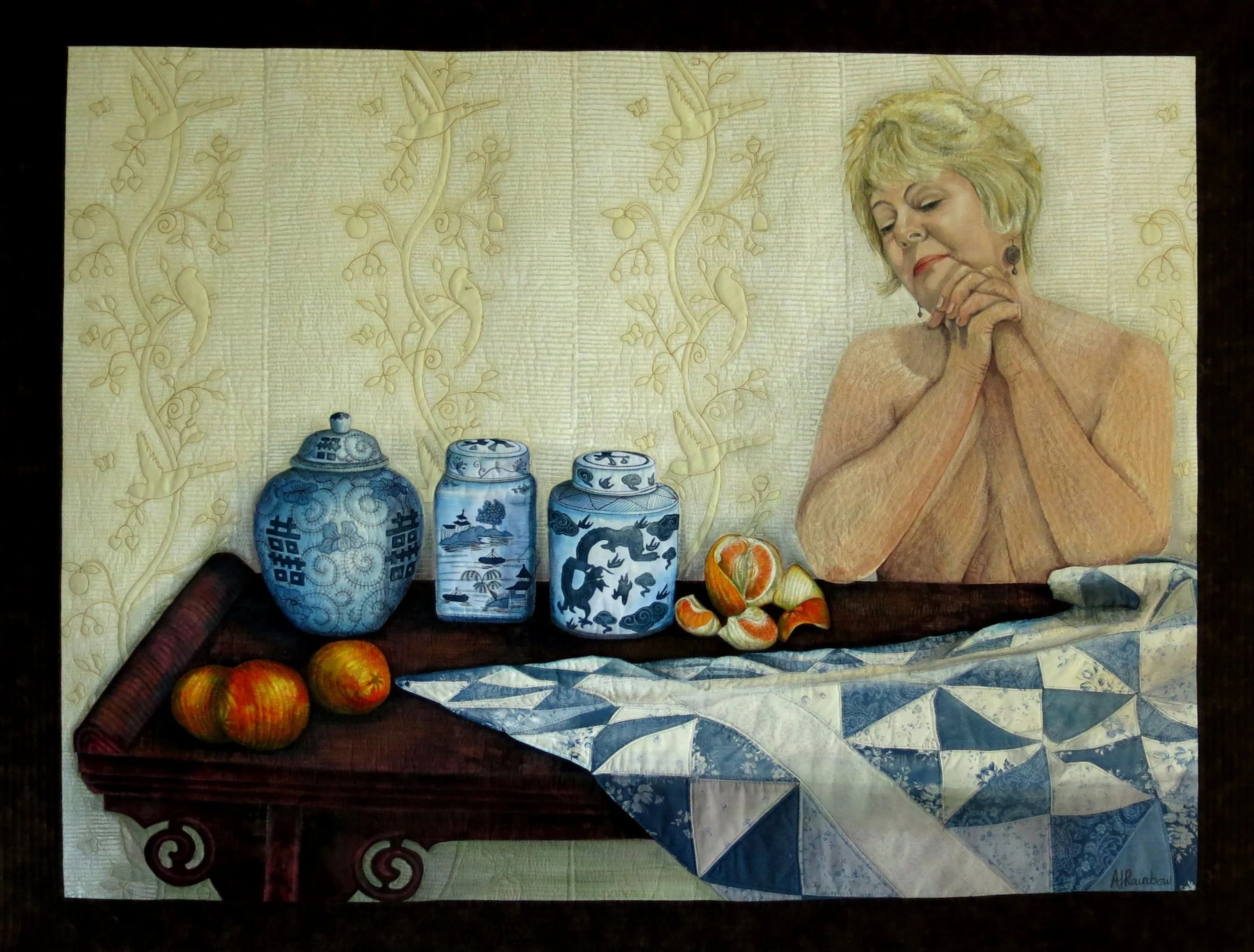 An art quilt by Annabel Rainbow featuring a woman with short blonde hair, clasping her hands, eyes closed, next to blue and white ceramic jars, a few oranges, and a blue quilted table cover on wooden table.