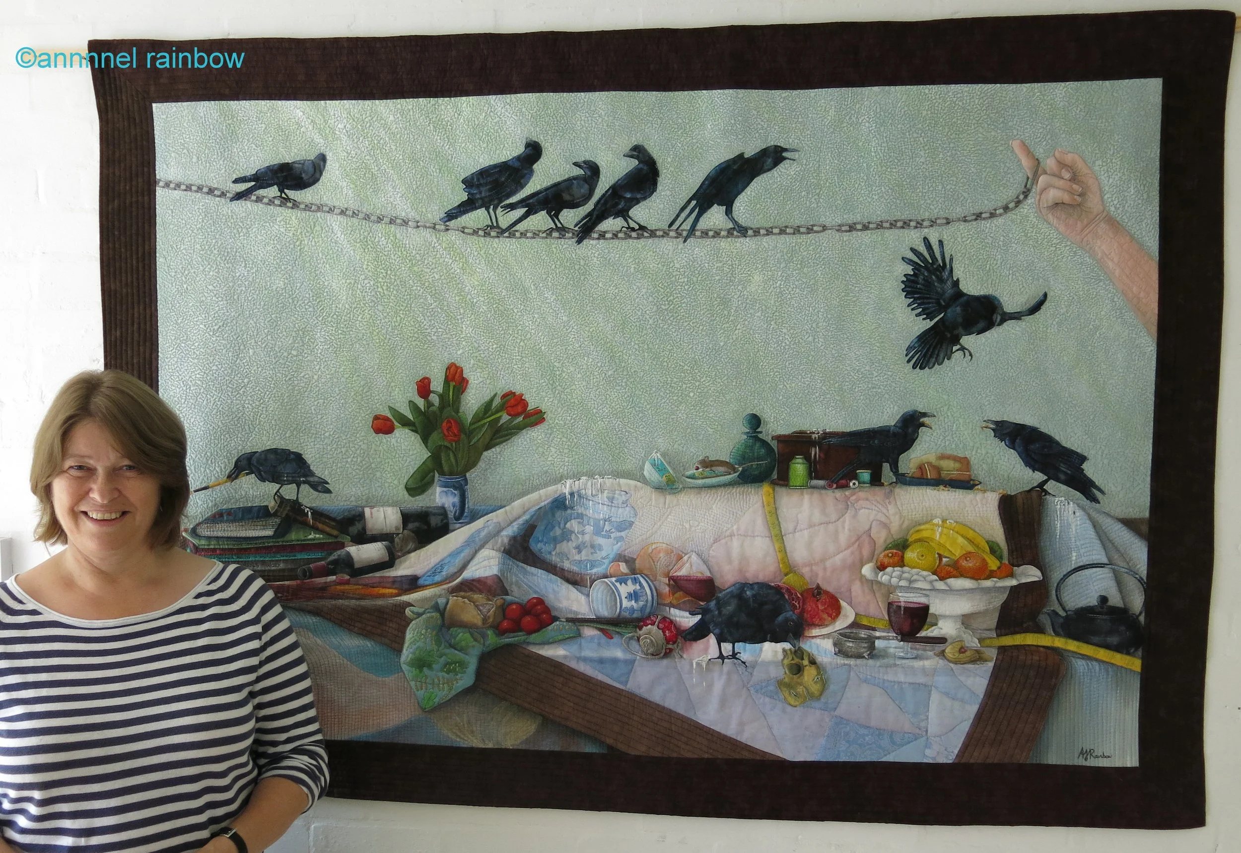 A Life Story quilt by Annabel Rainbow with her standing next to Murder, a fabric artwork depicting a domestic scene with crows, a vase of red tulips, and a table set with various food items and dishes.
