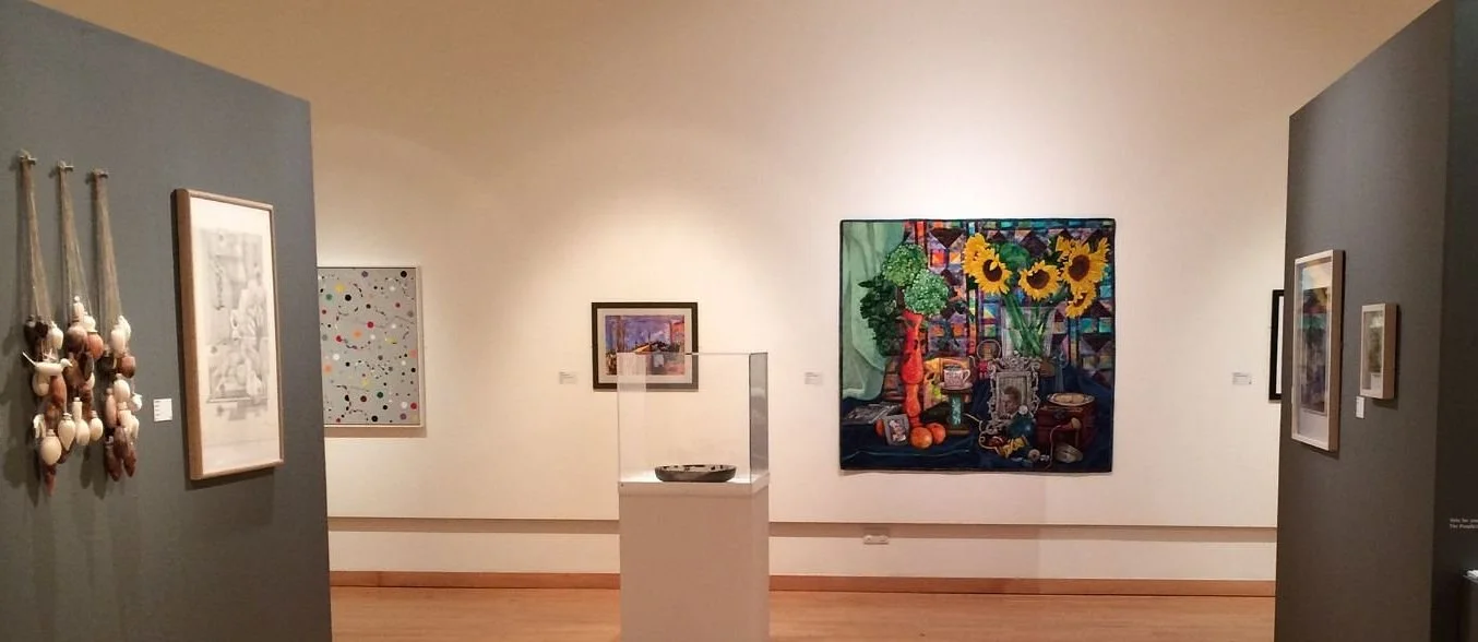 Award winning art quilt by Annabel Rainbow in a gallery interior with various artworks including paintings, a tapestry, and sculptures on display; central piece features sunflowers.
