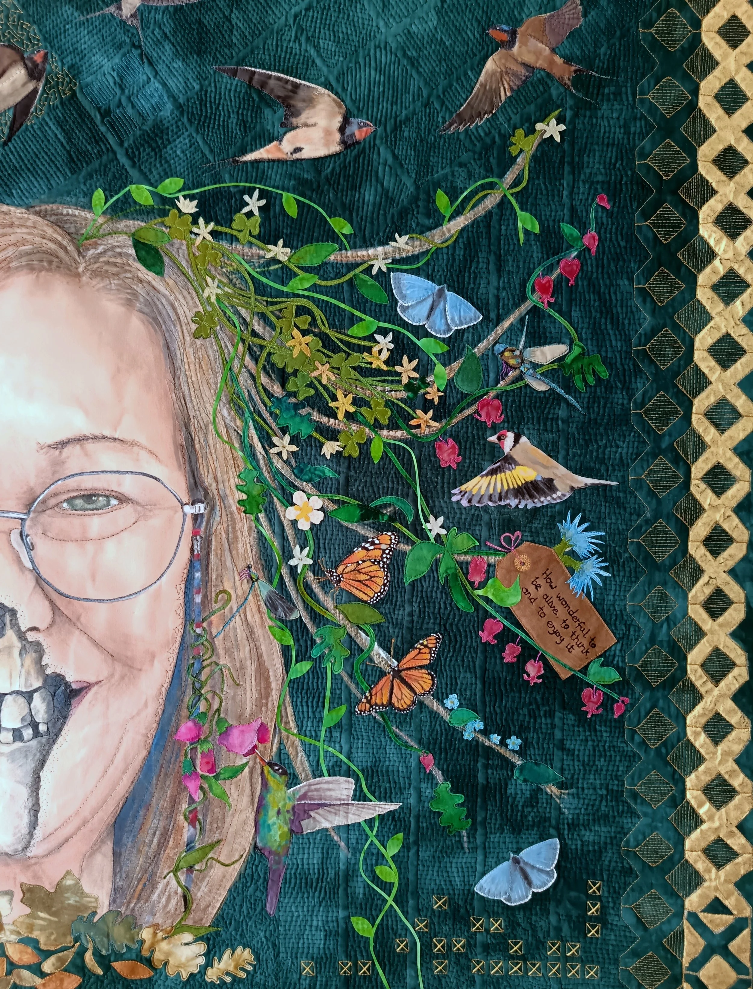 Artistic quilt featuring a half-face portrait of a person with glasses, surrounded by butterflies, birds, and foliage on a dark textured background. A note among the plants reads 'have wandered into a wild winter garden to enjoy unity.' The border is decorated with a gold geometric pattern.