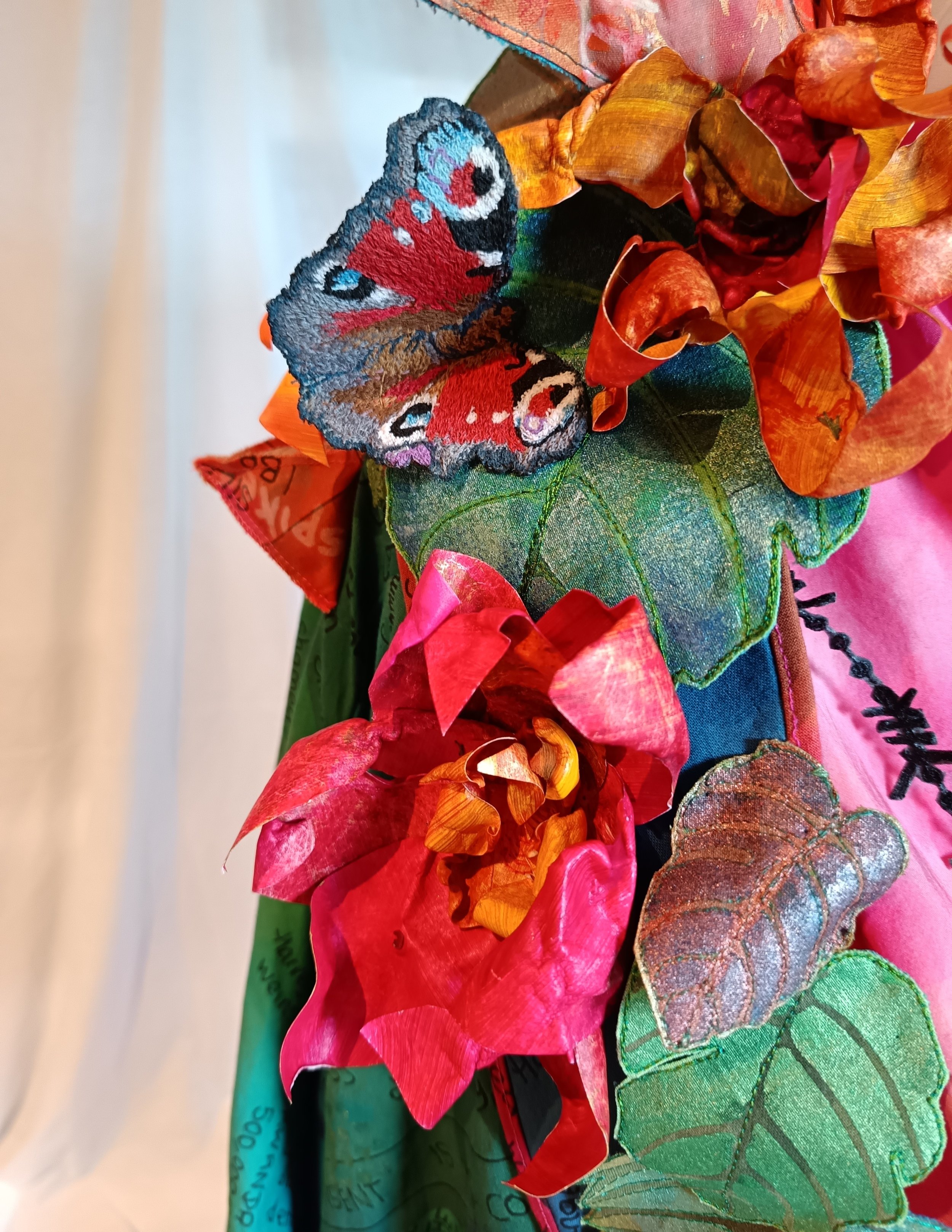 A decorative arrangement featuring a colorful cloth butterfly, artificial flowers, leaves, and fabric with embroidered text.