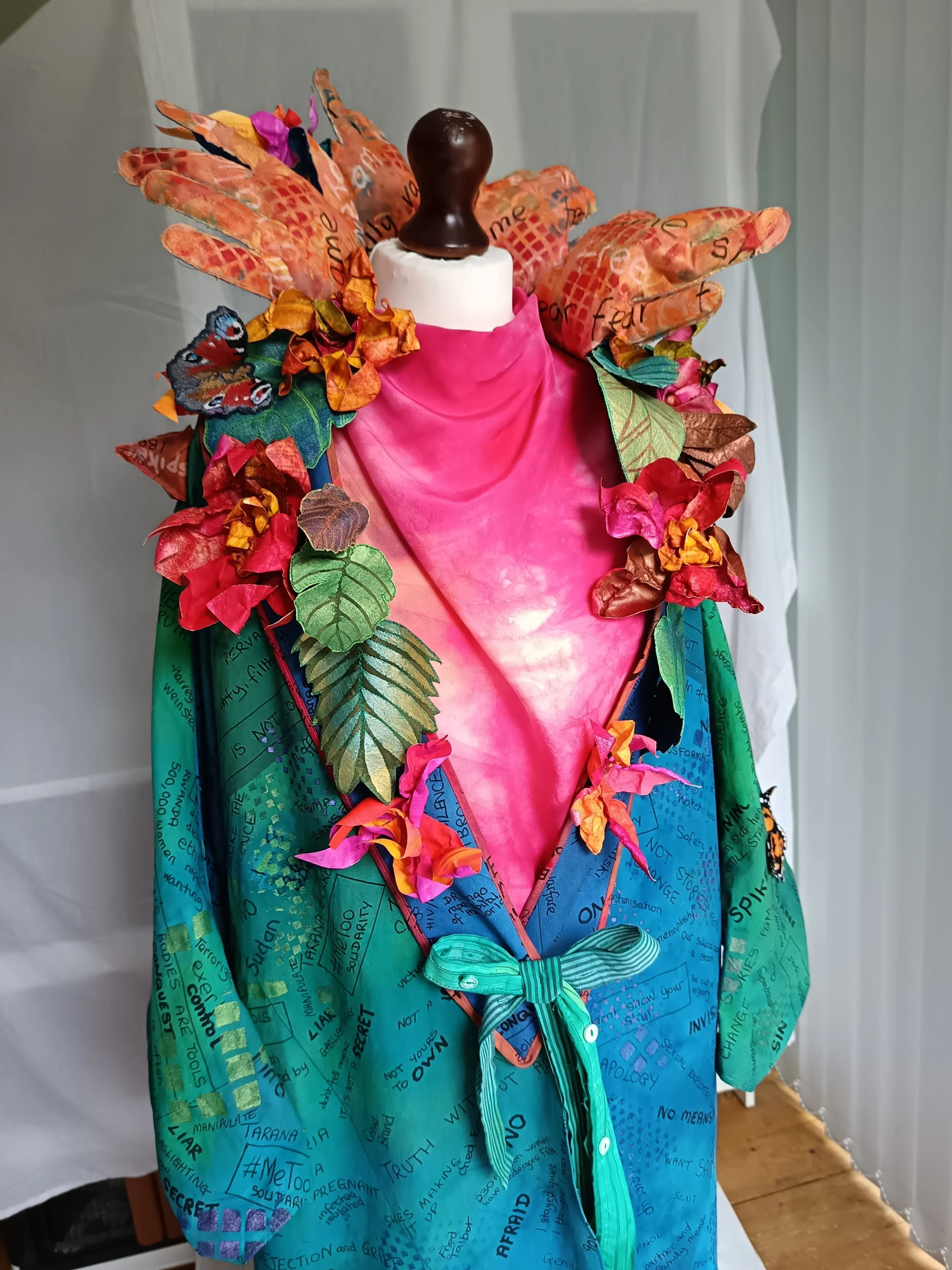 Colorful mixed-media dress featuring hand-painted text, fabric flowers, leaves, and butterflies, displayed on a mannequin.
