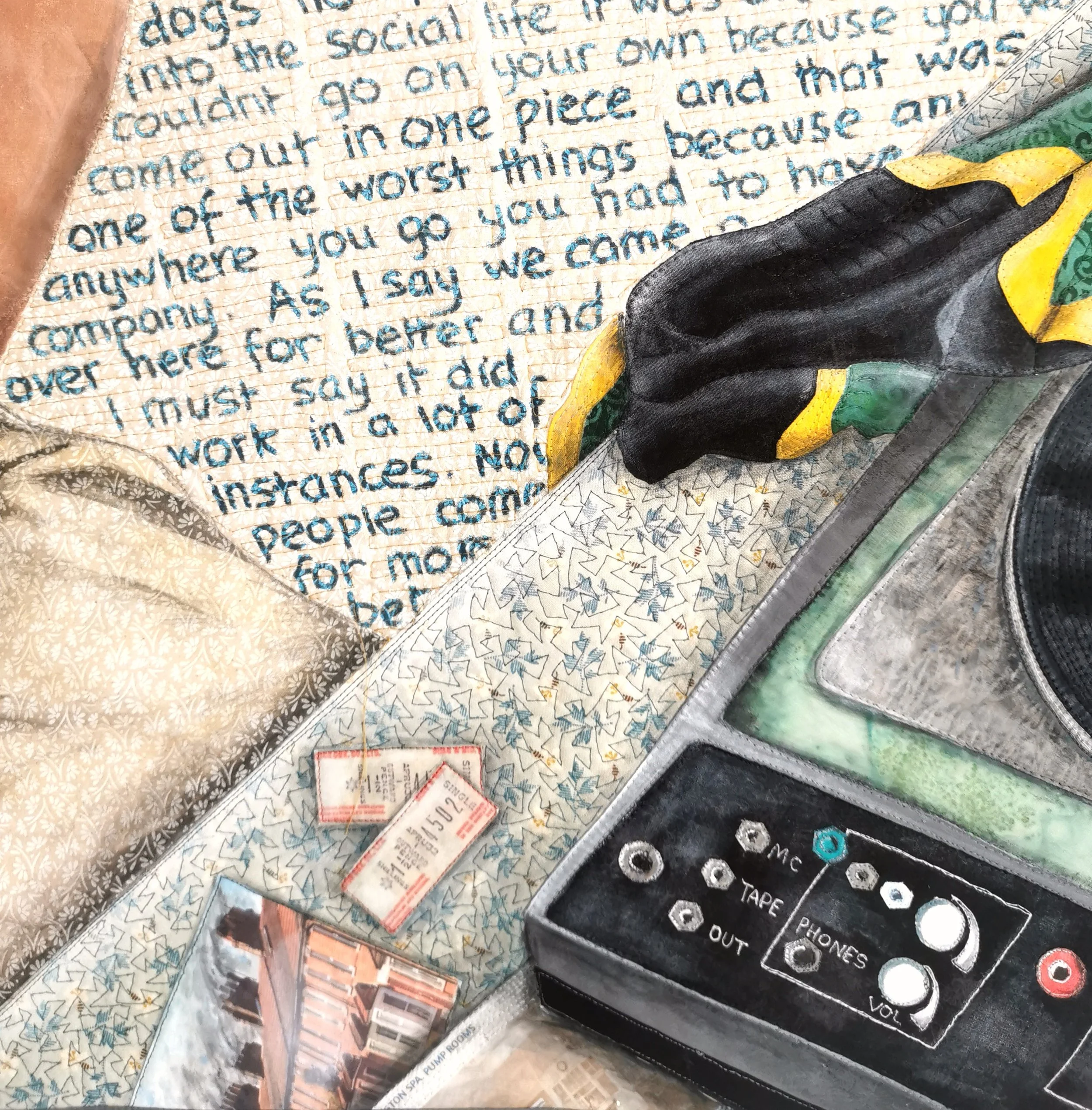 Close-up of a quilt with blue handwritten text, depicting a record player with buttons labeled MC, Tape, Phones, Out, and Vol. Nearby are concert tickets and a photograph featuring a cityscape. A yellow and black fabric is draped over the quilt.