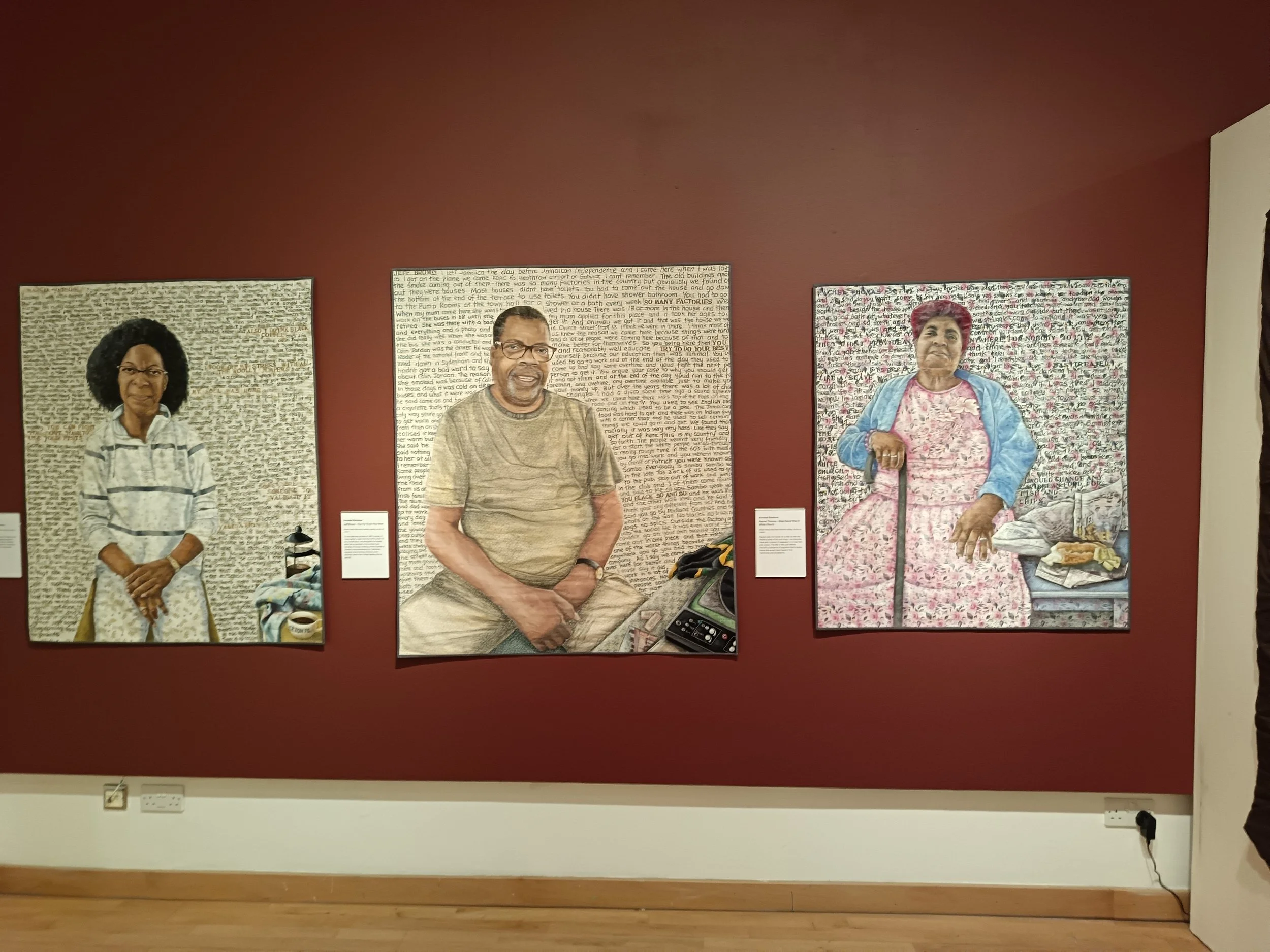Three portraits of elderly individuals displayed on a red wall in an art gallery, with informational plaques below each.