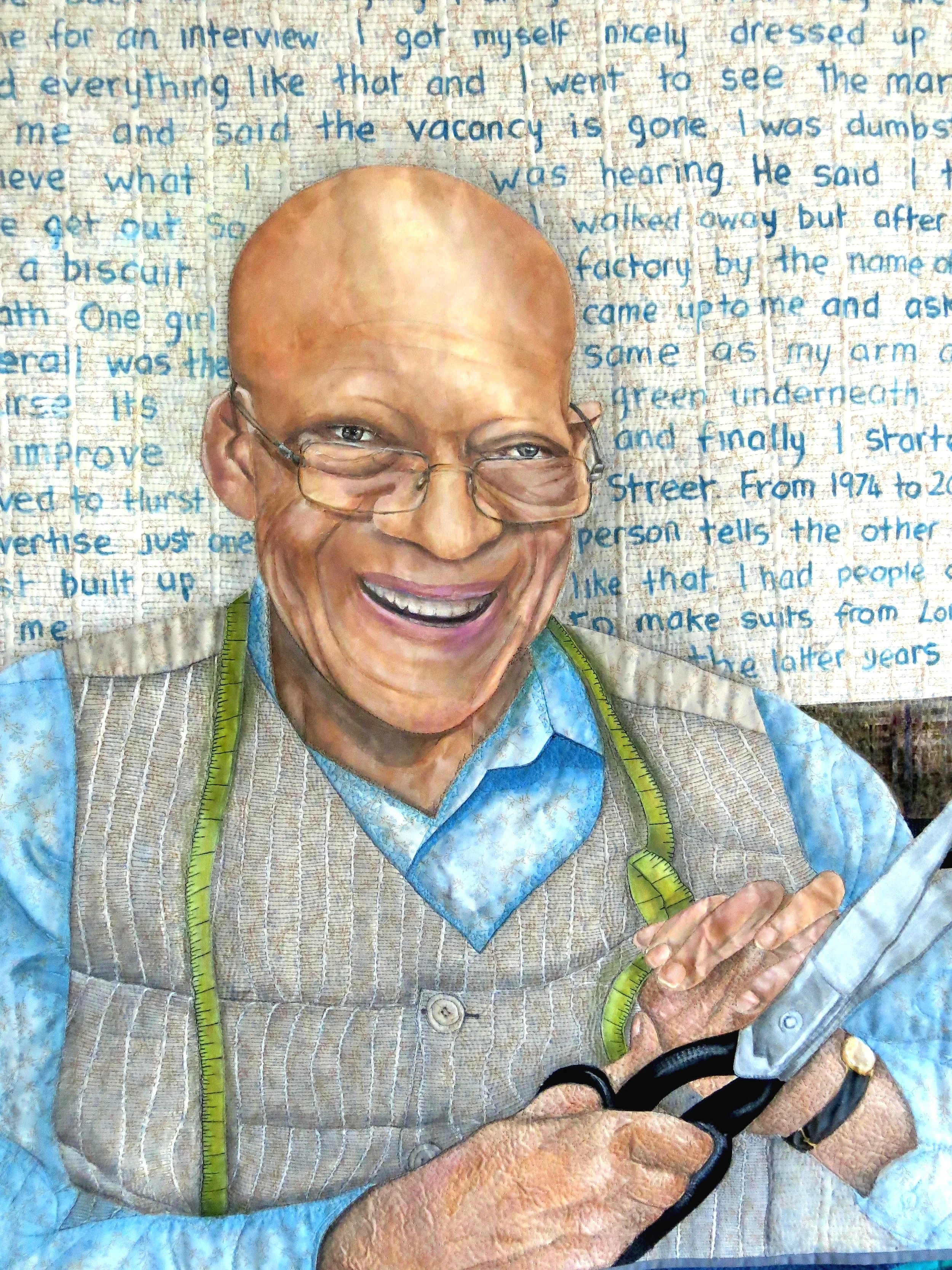 A textile artwork depicting a smiling man with glasses holding a pair of scissors. He's wearing a blue shirt and a striped vest with a measuring tape around his neck. The background is filled with handwritten text.