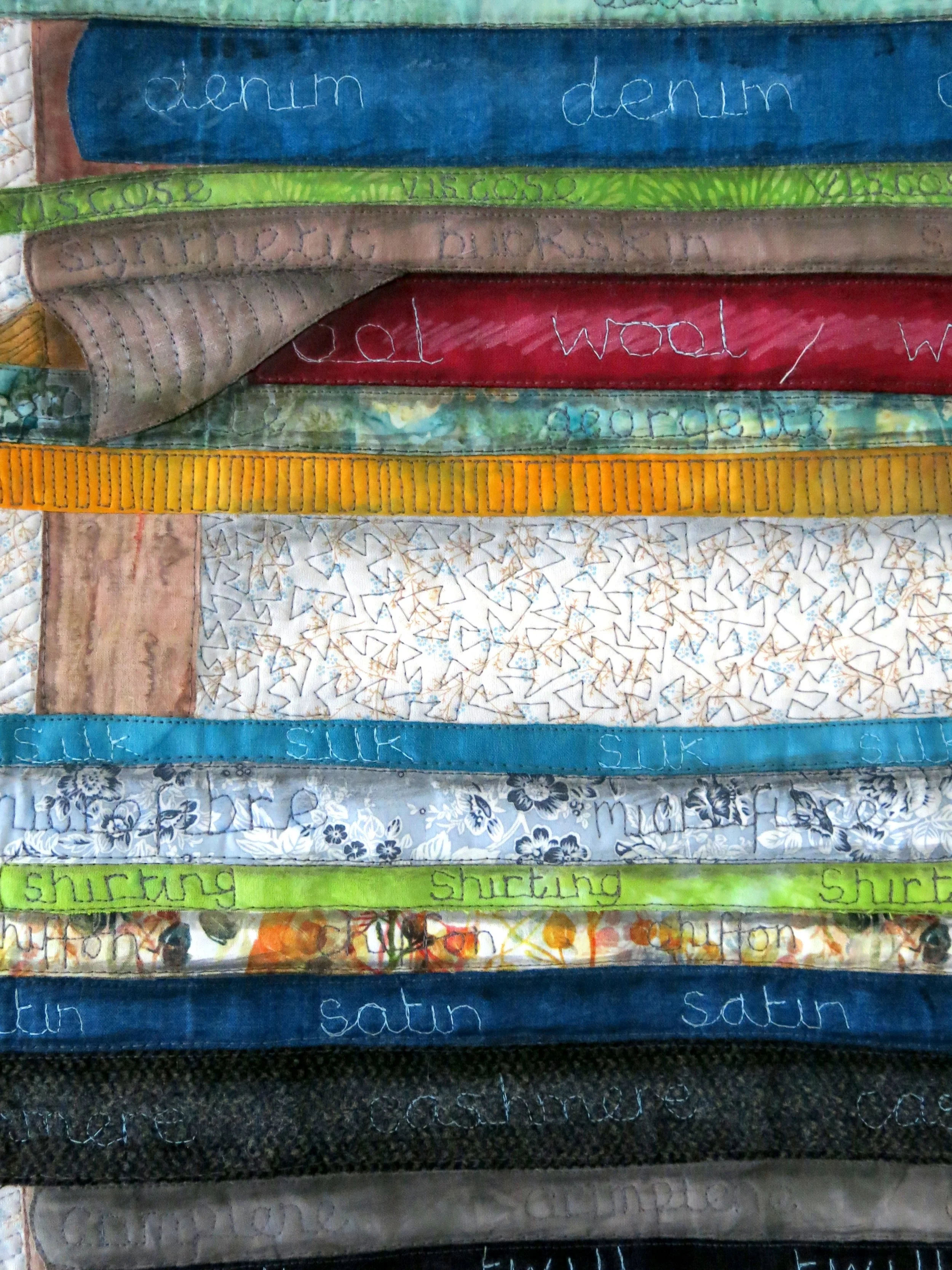 Close-up of a fabric art quilt featuring various textured and colored strips with text labels such as denim, wool, silk, and satin stitched on them.