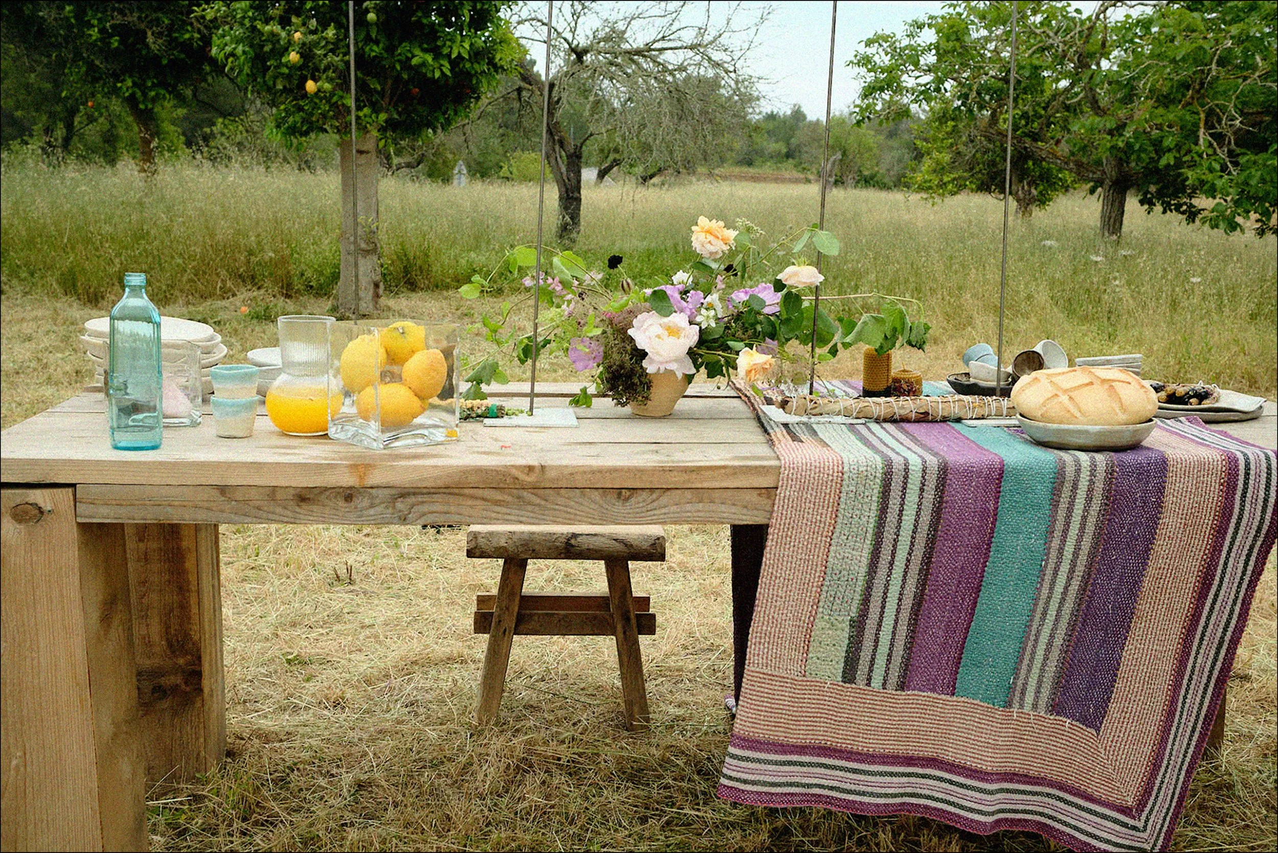 Outdoor table set with a colorful striped tablecloth, glass bottles, bowls, a loaf of bread, flowers in a vase, and lemons in a glass bowl, with trees and grassy field in the background.