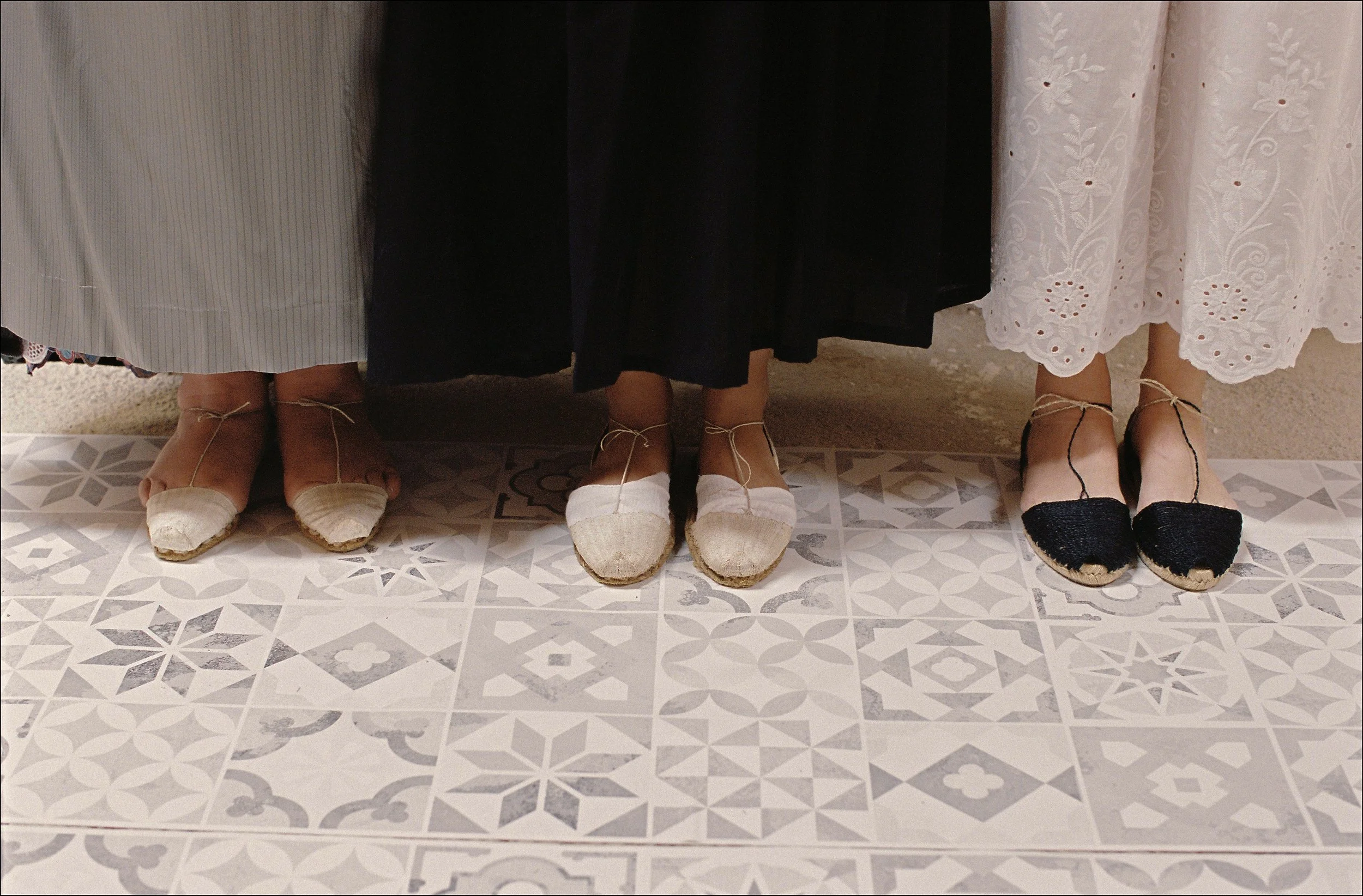The lower halves of three women standing side by side, wearing long skirts and espadrille shoes, traditional shoewear of the Balearic Islands, with patterned floor tiles beneath their feet.