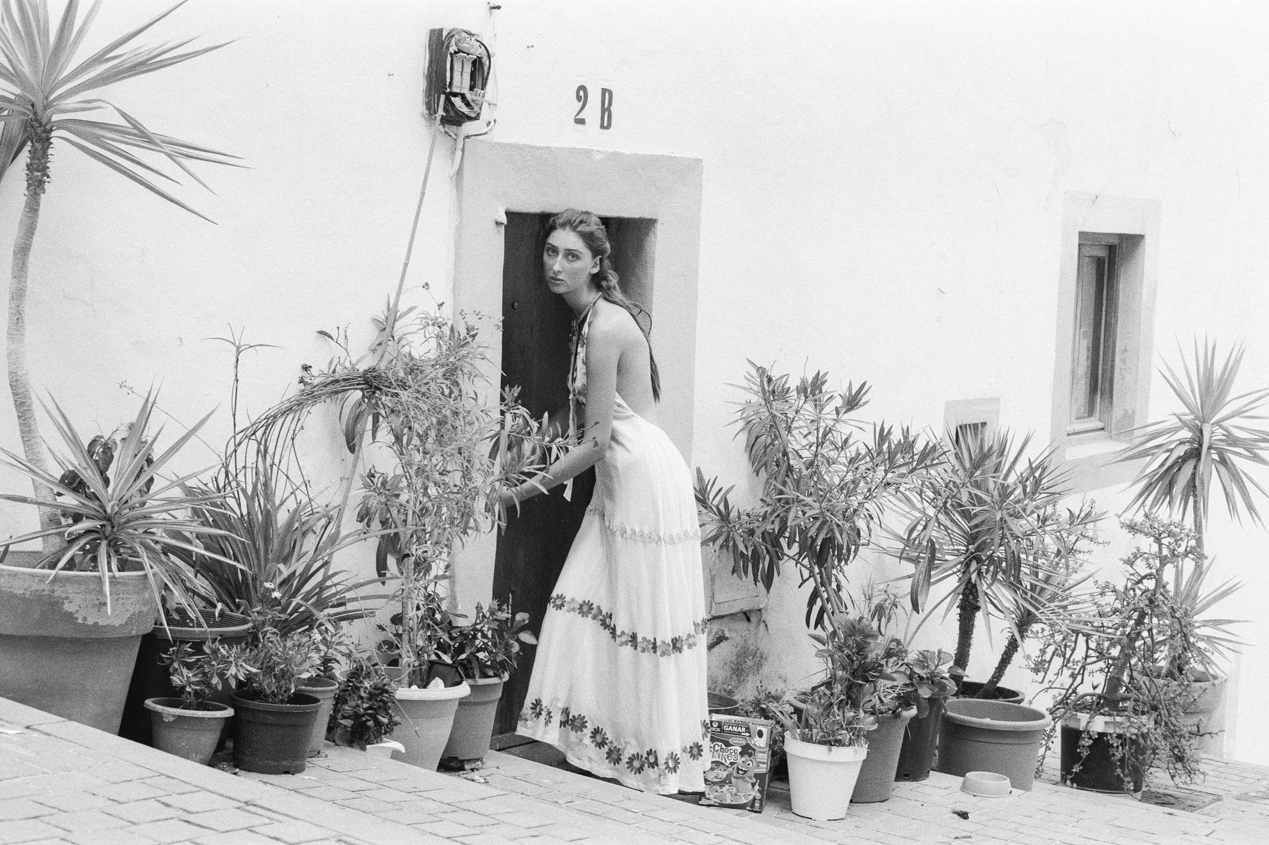 A woman wearing a Balearic artisan made patterned dress stands among potted plants on a patio. The wall behind her has the number '2B' and a small window. There are plants and gardening items around her.