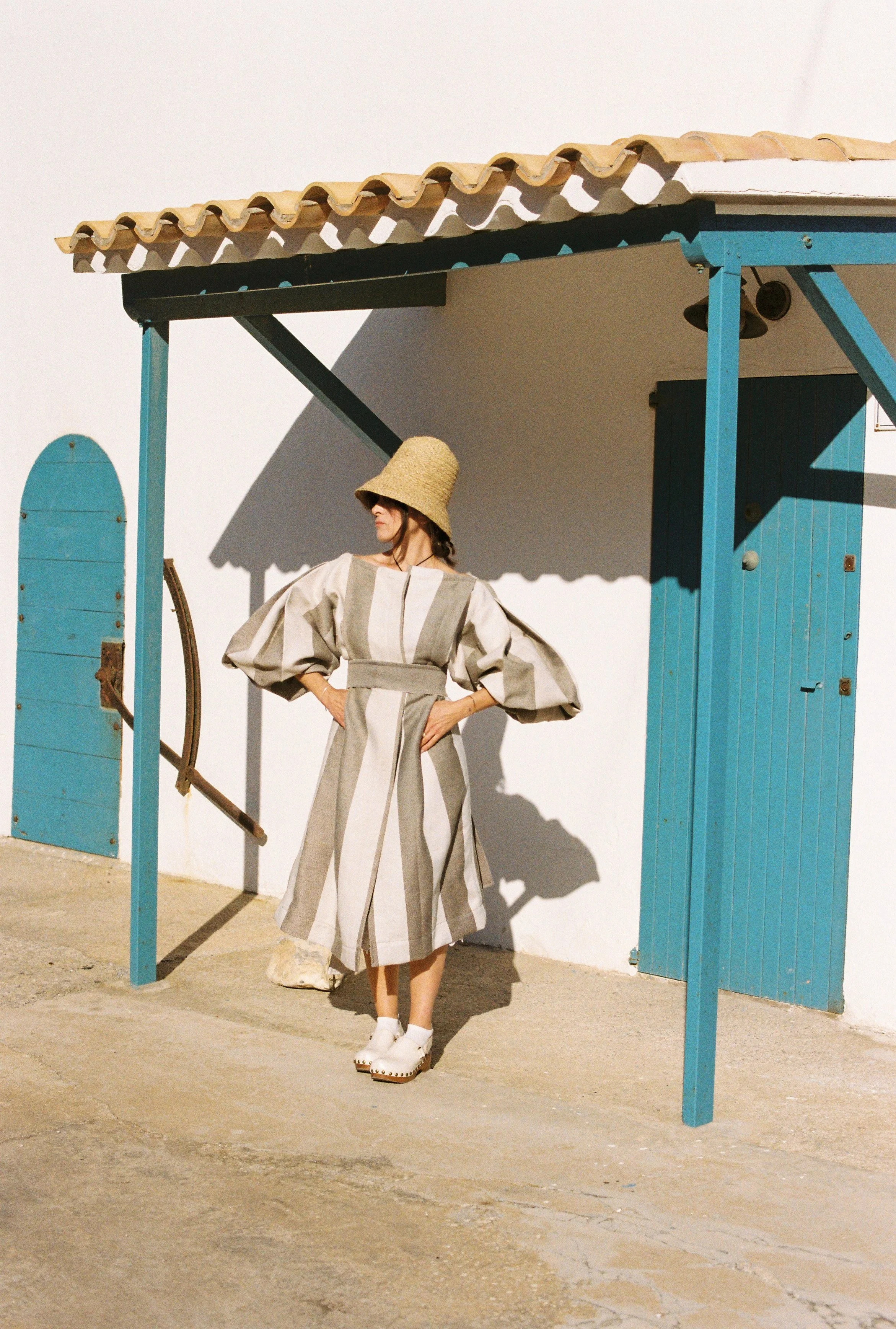 Mariola Escofet, The Balear edit's founder, wearing a contemporary design by an Ibizan designer: a large straw hat, a beige and gray striped dress with puffy sleeves, white shoes; standing outdoors in an authentic Ibizan setting.