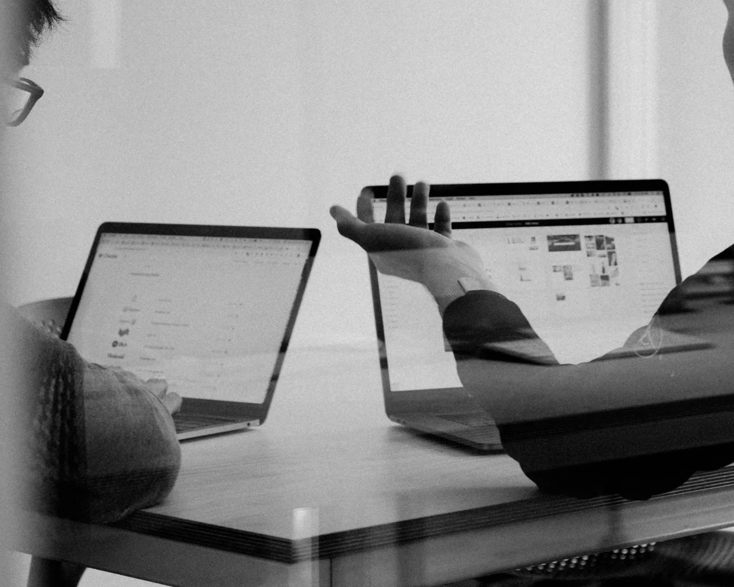 Deux personnes utilisant des ordinateurs portables sur une table en réunion, avec un fond blanc, en noir et blanc.
