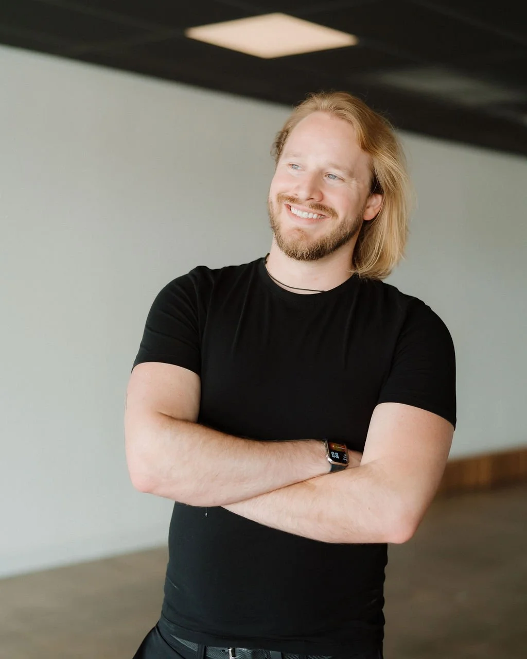 A young man with long, blond hair, a beard, and a black T-shirt smiling with arms crossed in a modern indoor setting.
