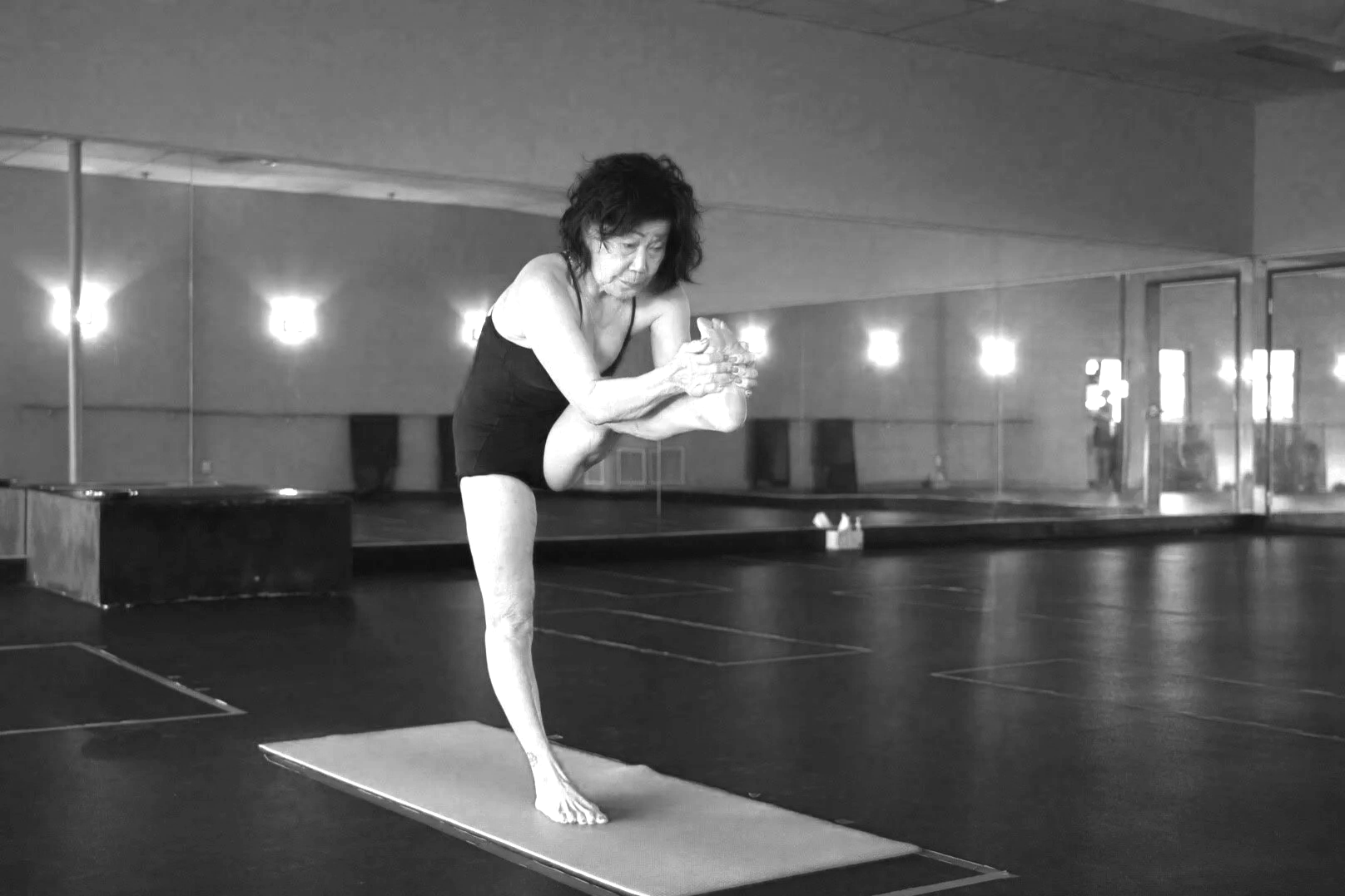 An elderly woman practicing yoga indoors on a mat, balancing on one leg with her other leg bent and held in front of her, in a room with mirrored walls and soft lighting.