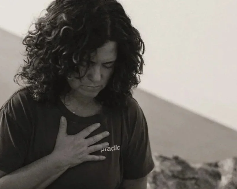 A woman with curly hair, eyes closed, holding her chest, appearing to experience emotional pain or distress.