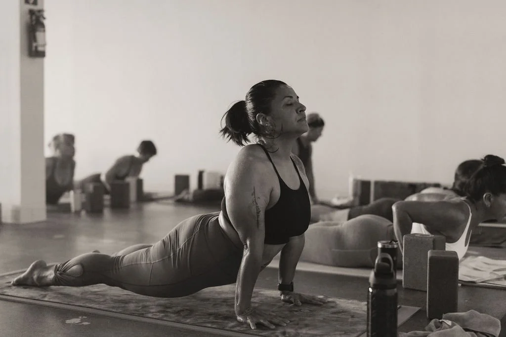 A woman practicing yoga in a studio is doing the upward dog pose, with other participants in the background also performing yoga poses.
