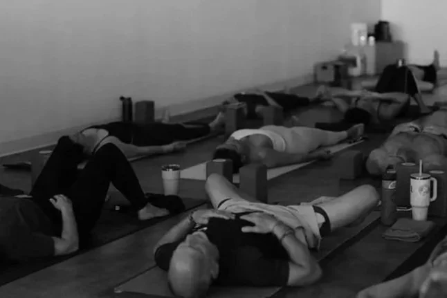 People practicing yoga or meditation in a dimly lit studio, lying on mats with drinks and personal items nearby.