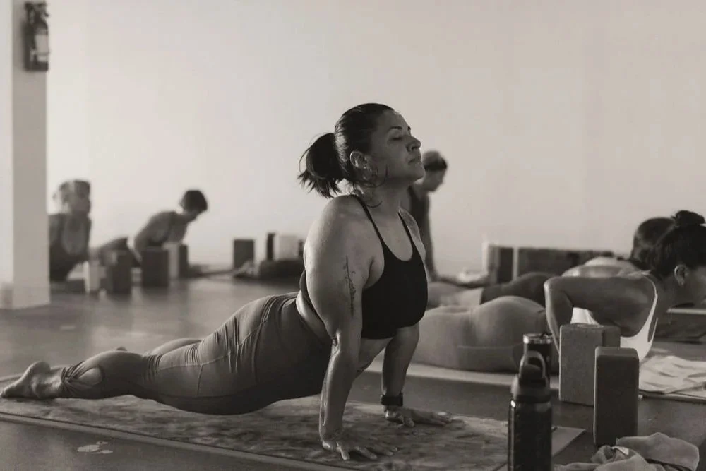 A woman practicing yoga in a classroom, in a yoga pose with her body parallel to the ground and arms extended, surrounded by other women in similar positions.