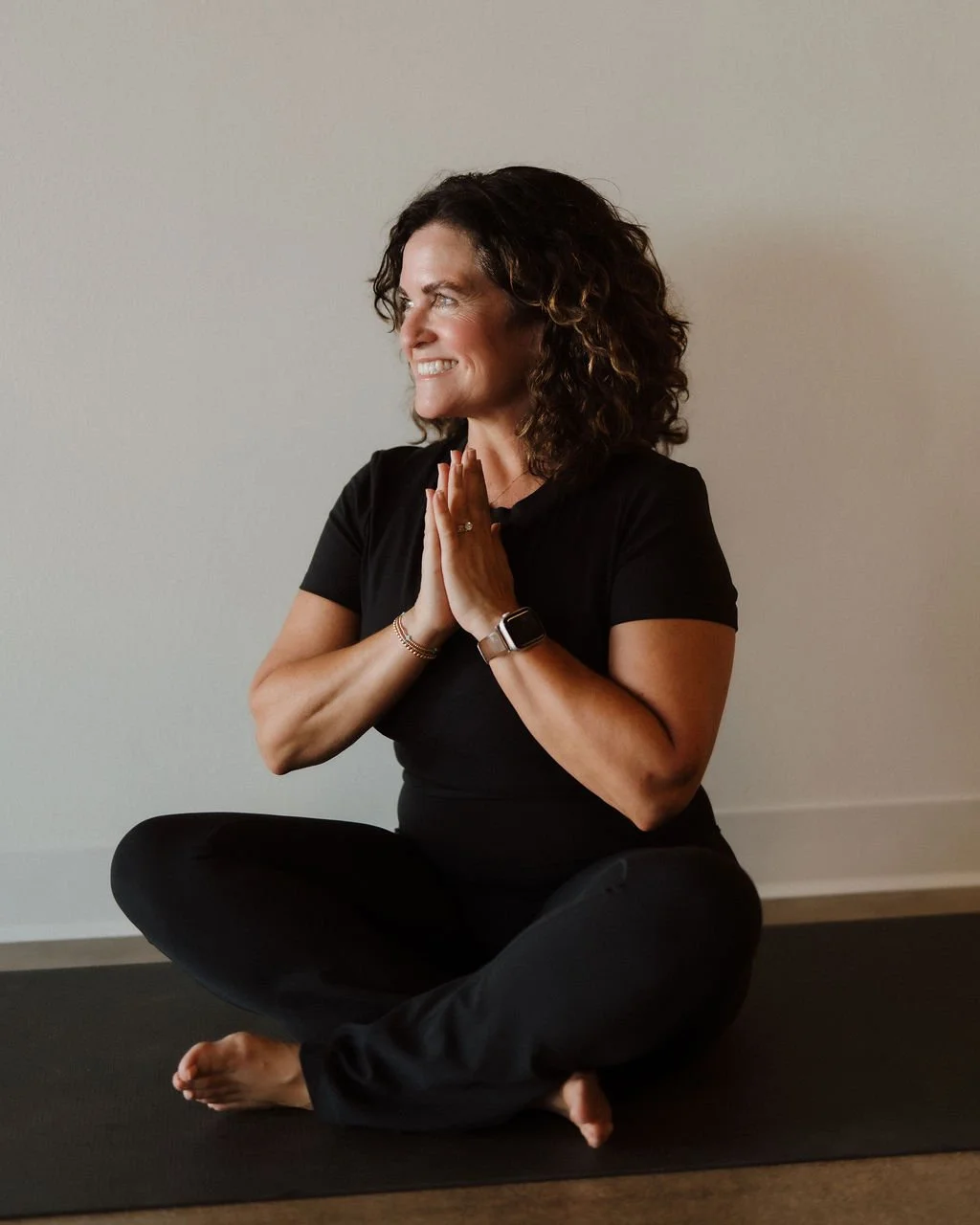 Woman practicing yoga or meditating indoors, sitting cross-legged on a black yoga mat, with hands together in prayer position, smiling and looking to the side.